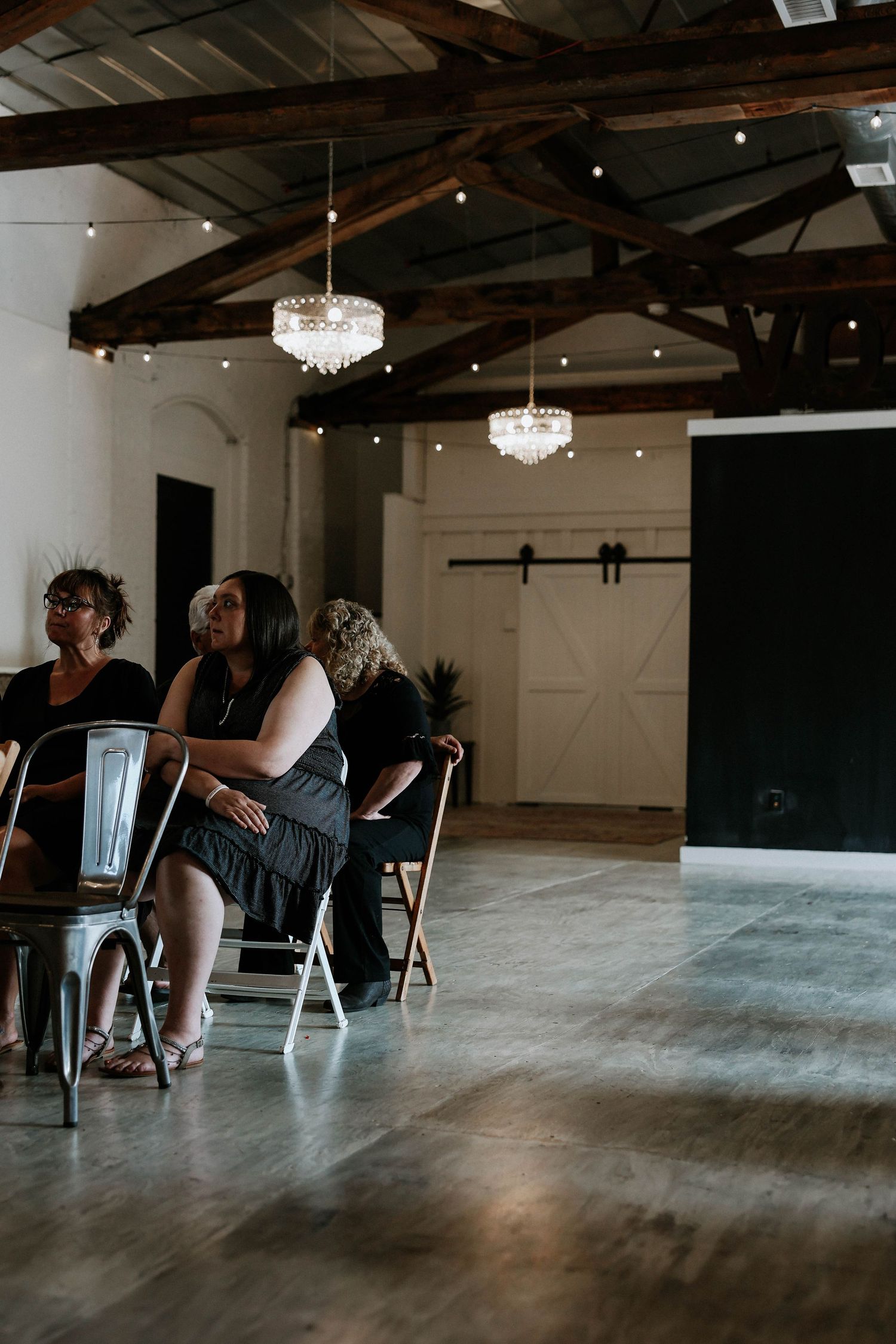 Guests seated in chairs at an elegant rustic venue with exposed wood beams and crystal chandeliers.