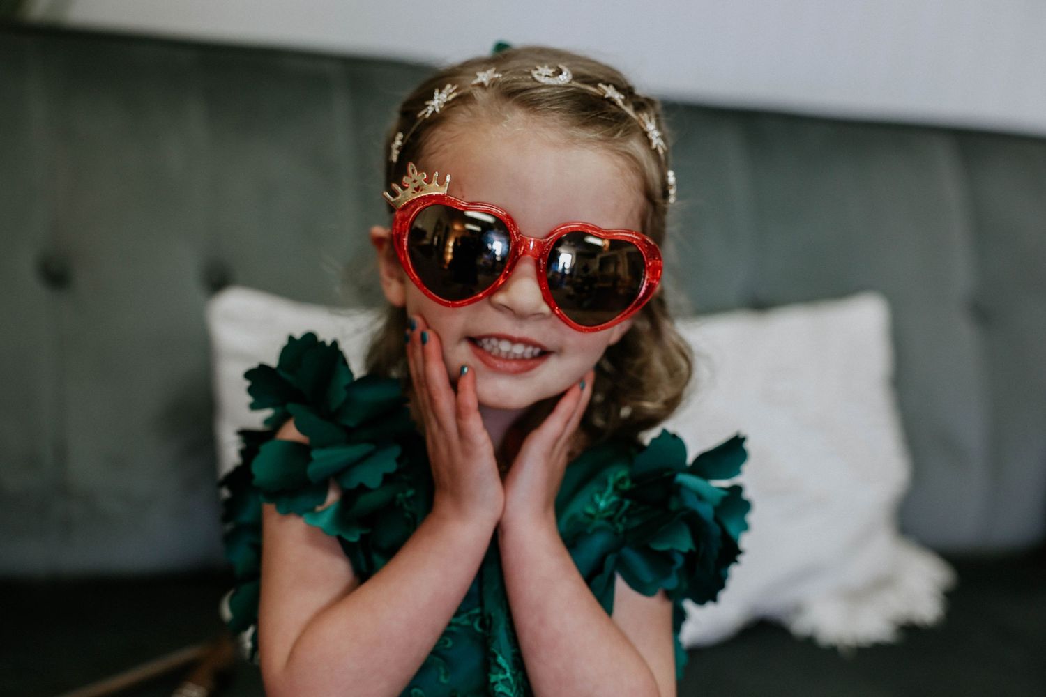 Adorable little girl in green dress wearing red heart-shaped sunglasses and gold crown headband smiling.