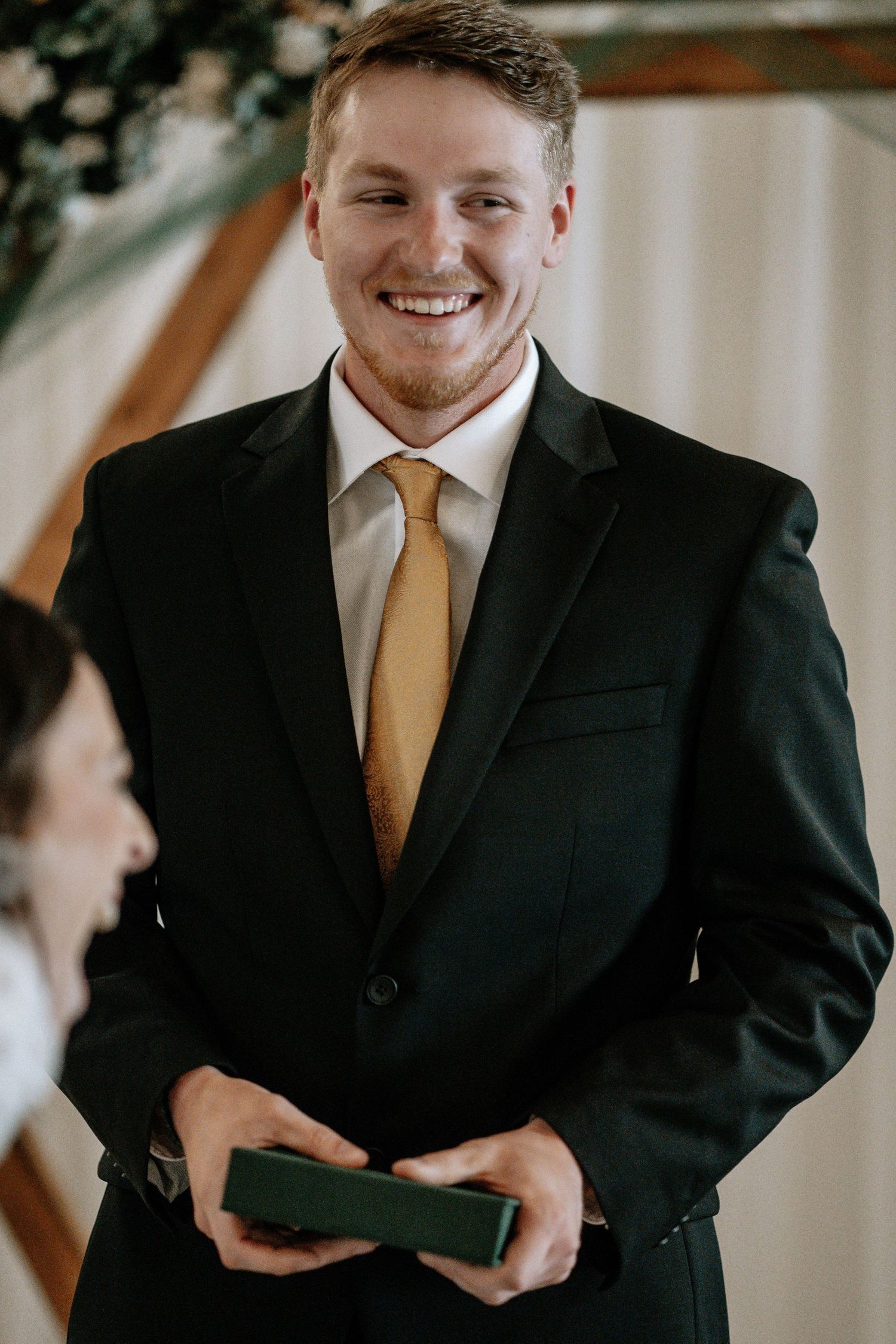 Smiling groom in black suit and gold tie holding a green ring box at a wedding ceremony.