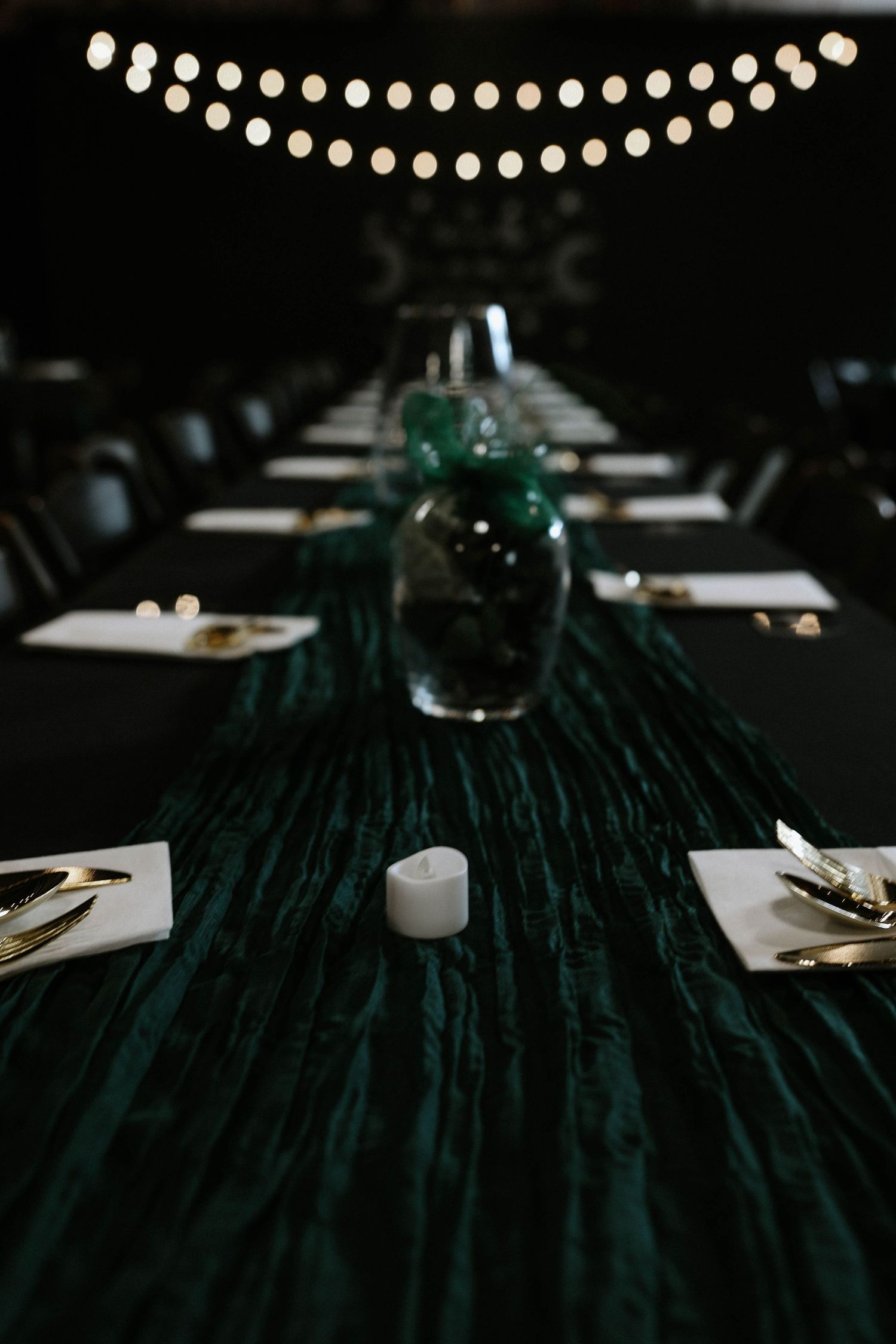 Elegant dark dining table with emerald green velvet runner, gold cutlery, white candles, and string lights above.