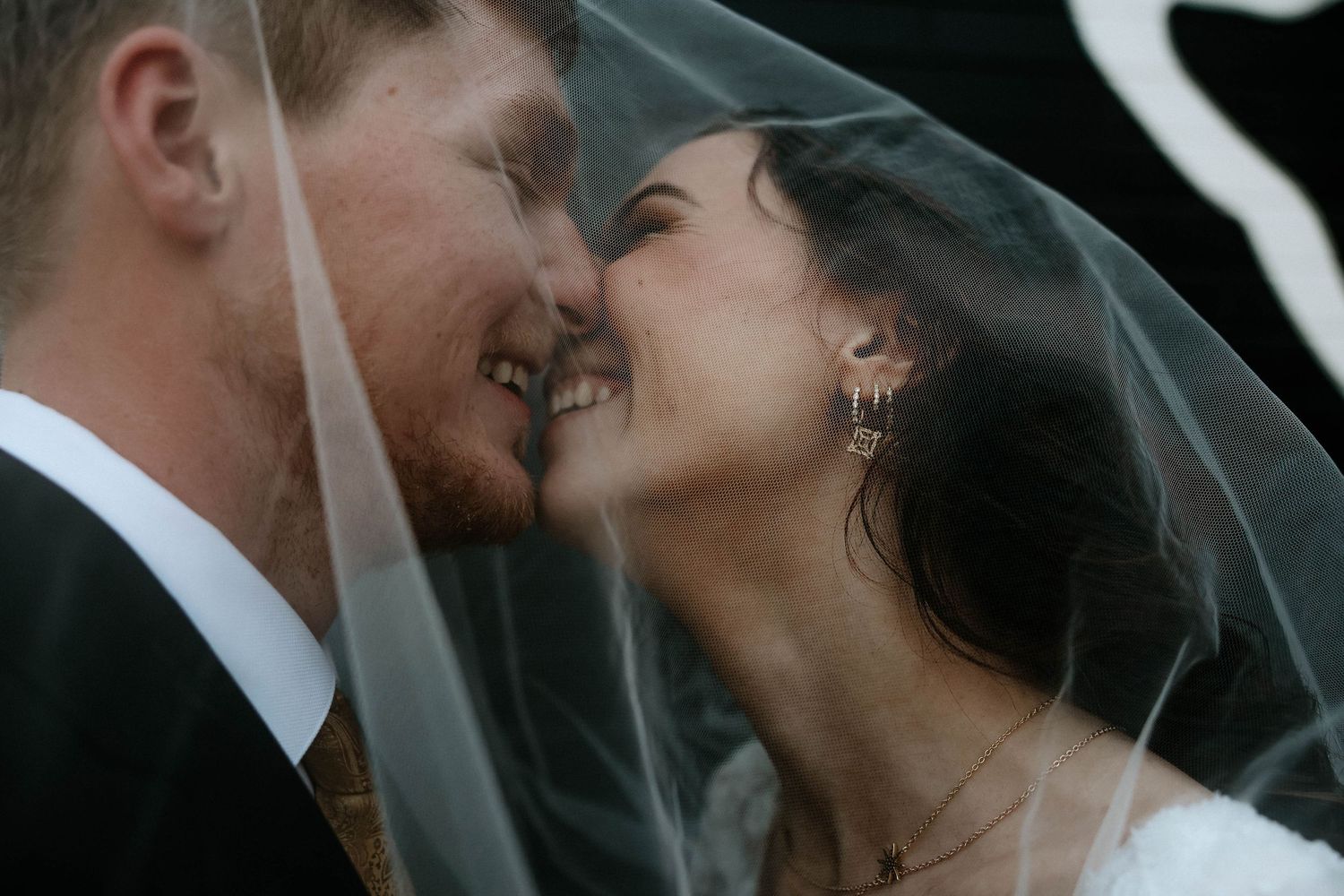 A bride and groom share a romantic moment under a wedding veil, smiling and touching foreheads on their wedding day.