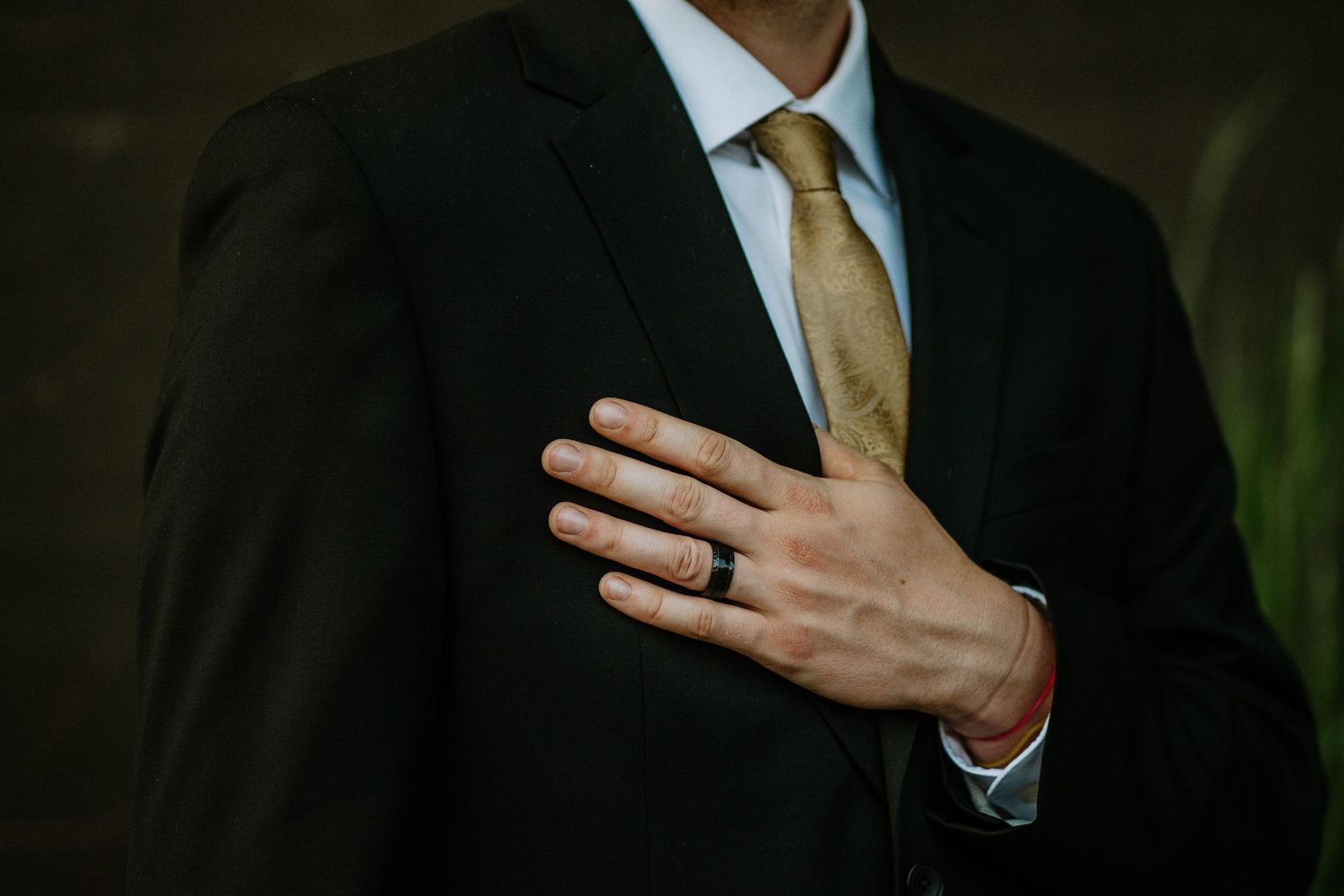Man in black suit with gold tie placing hand on chest, wearing a black wedding band ring.