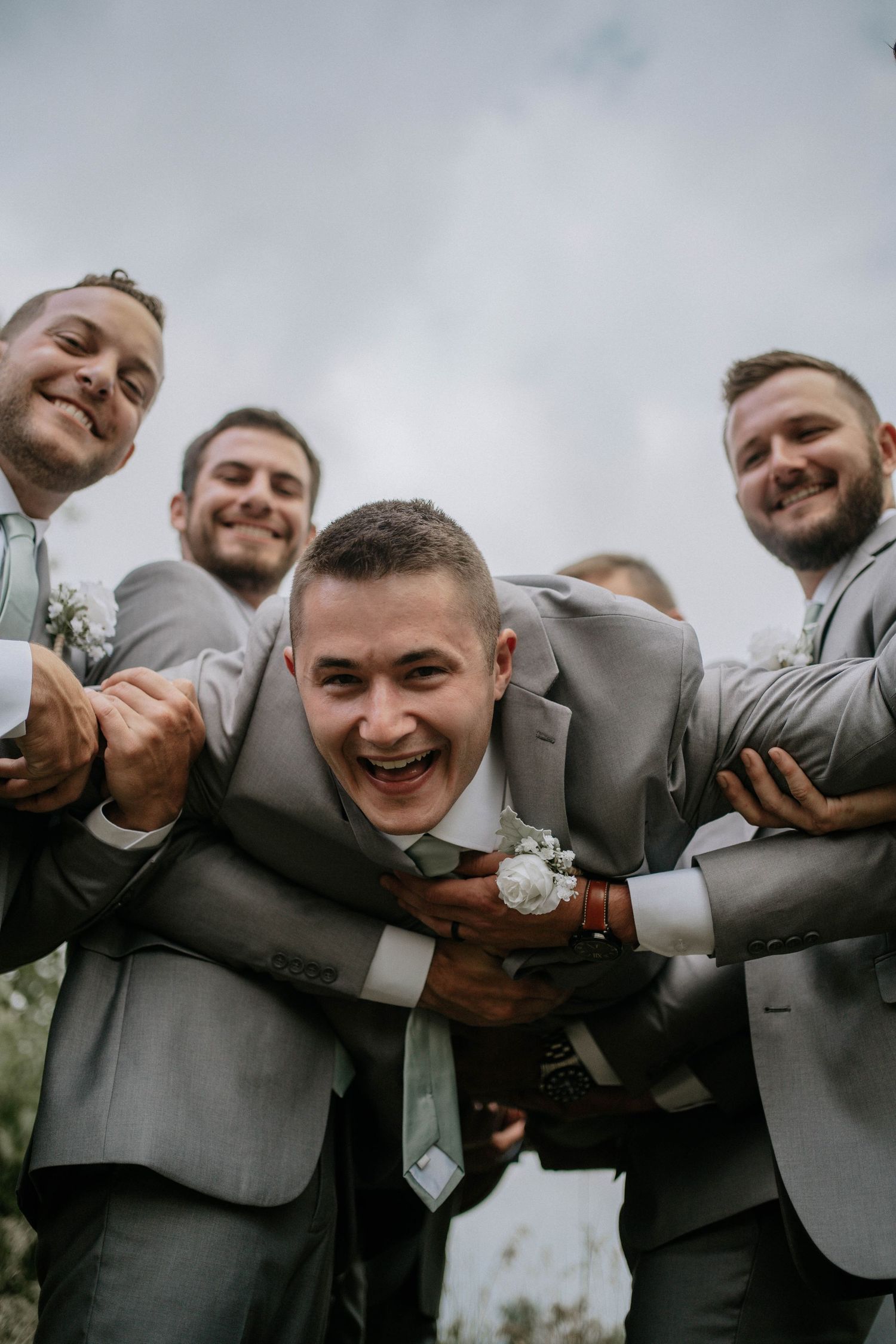 Groomsmen in gray suits playfully hugging the groom outdoors at a wedding celebration.