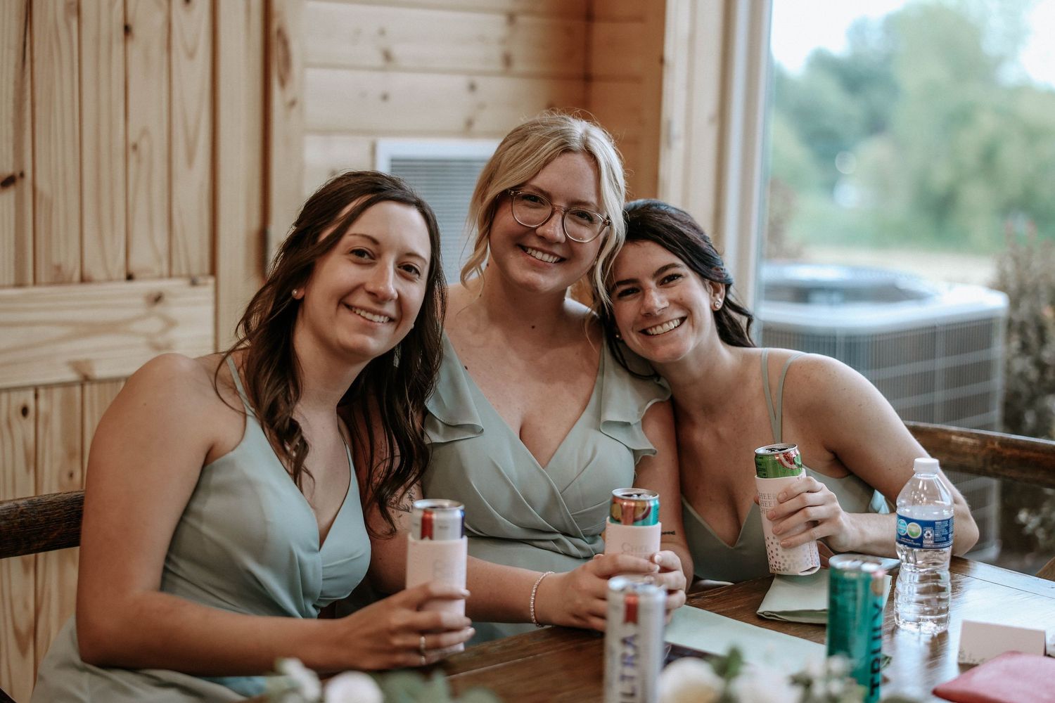 Three bridesmaids in sage green dresses smiling and holding drinks at a rustic wooden wedding reception venue.