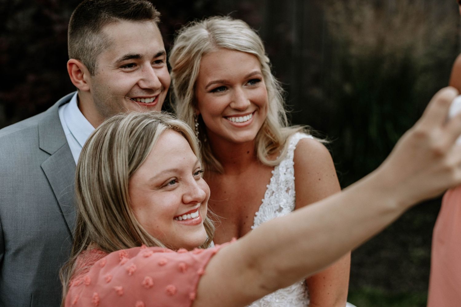 Bride in lace wedding dress takes a selfie with two smiling guests at an outdoor wedding celebration.