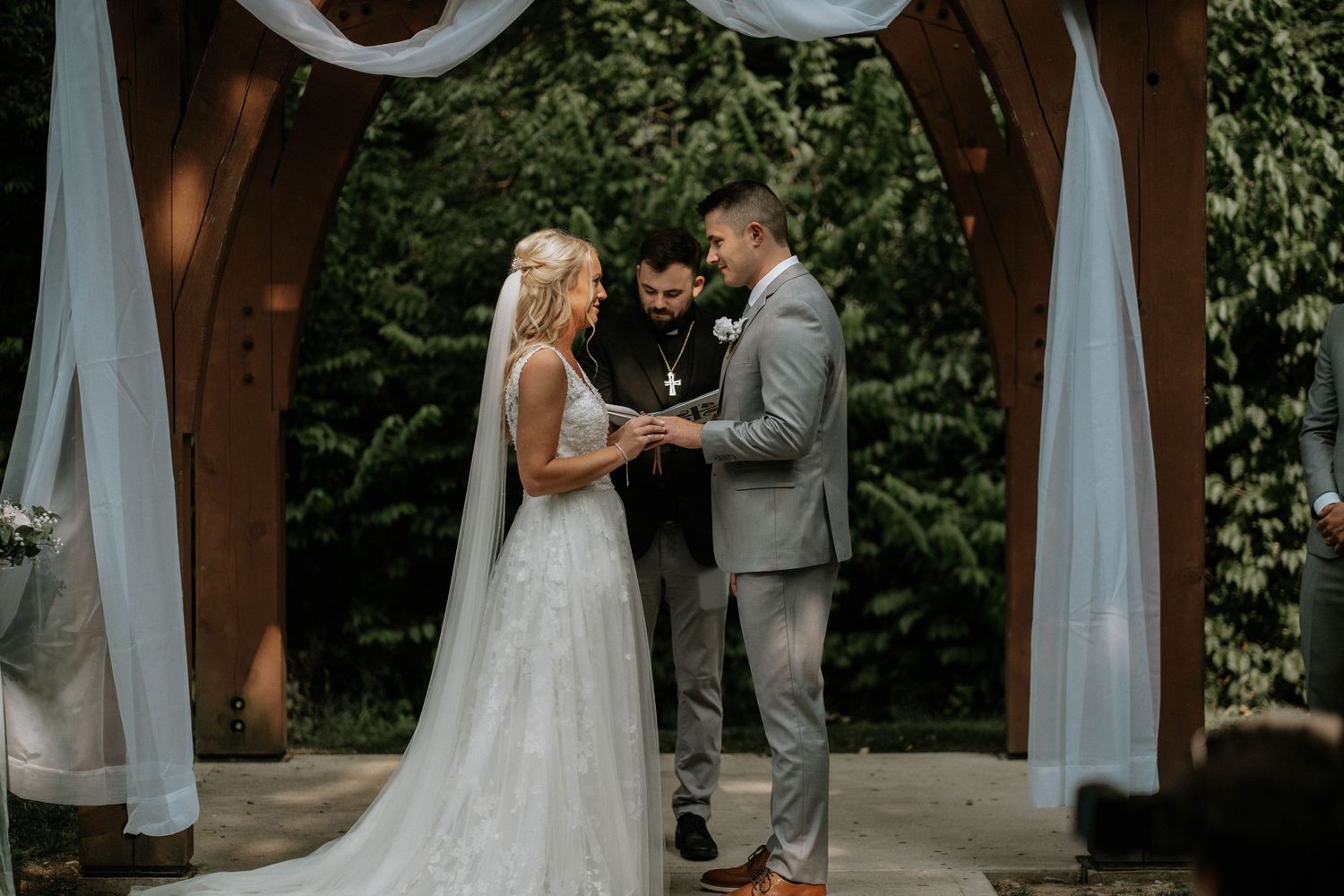 Bride and groom exchange vows under a wooden arch with white draping at an outdoor wedding ceremony.