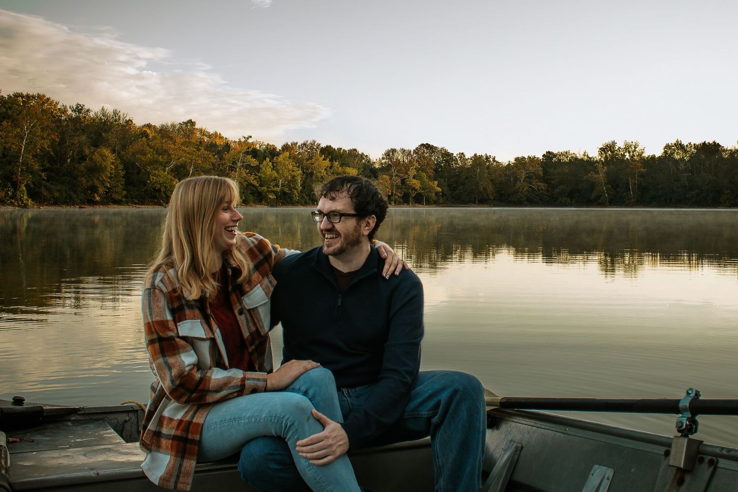 A couple sits together on a boat dock by a misty lake at sunrise, smiling at each other in Cincinnati, Ohio