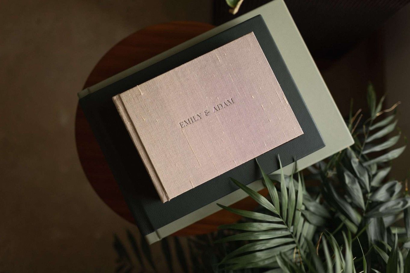 Elegant pink linen wedding album with embossed names resting on a dark surface with tropical leaves in Cincinnati, Ohio