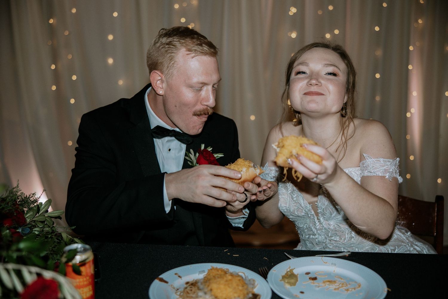 Bride and groom in wedding attire laugh together while eating food at their reception dinner table with fairy lights.