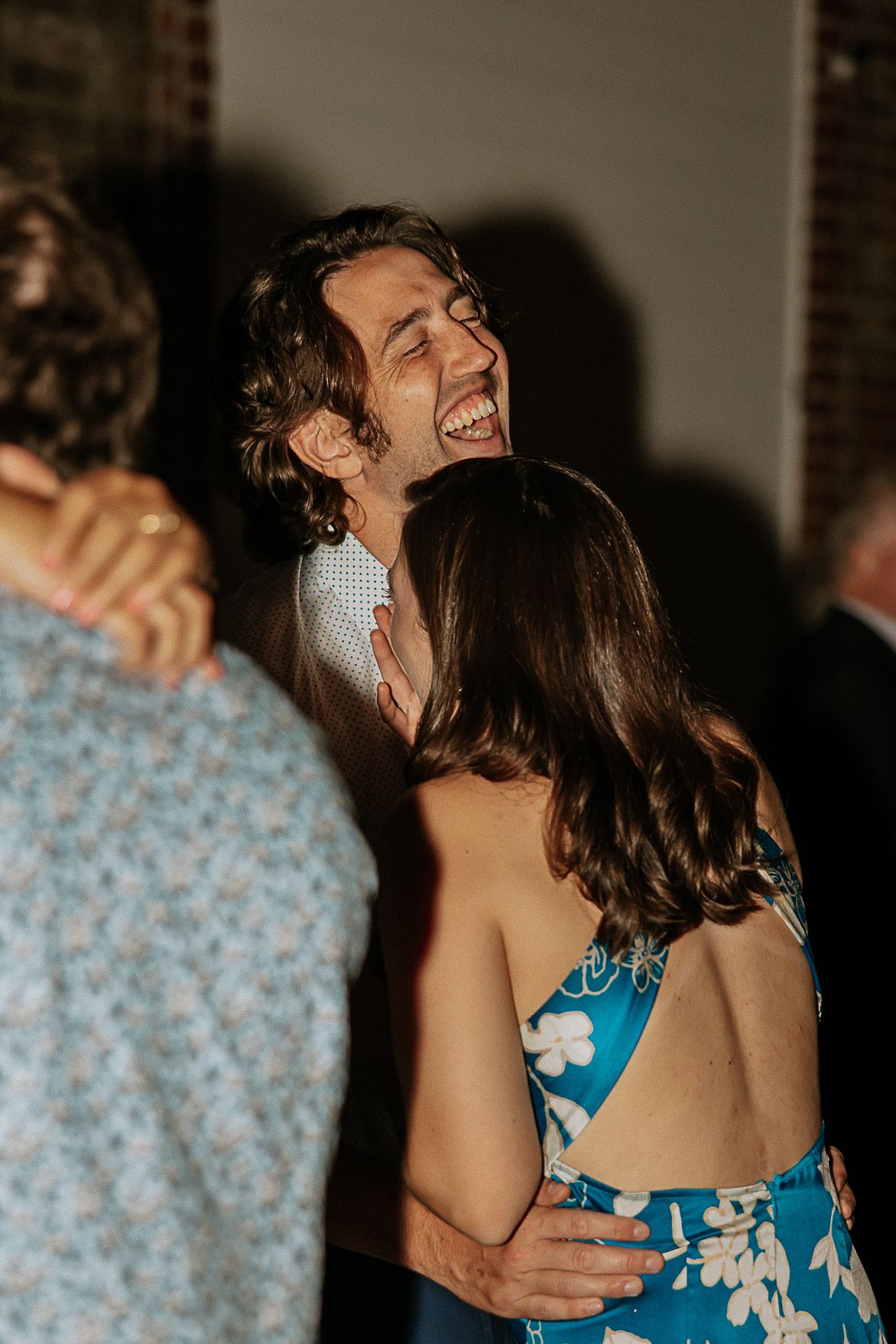 A man laughs joyfully while embracing a woman in a floral dress at a wedding reception.