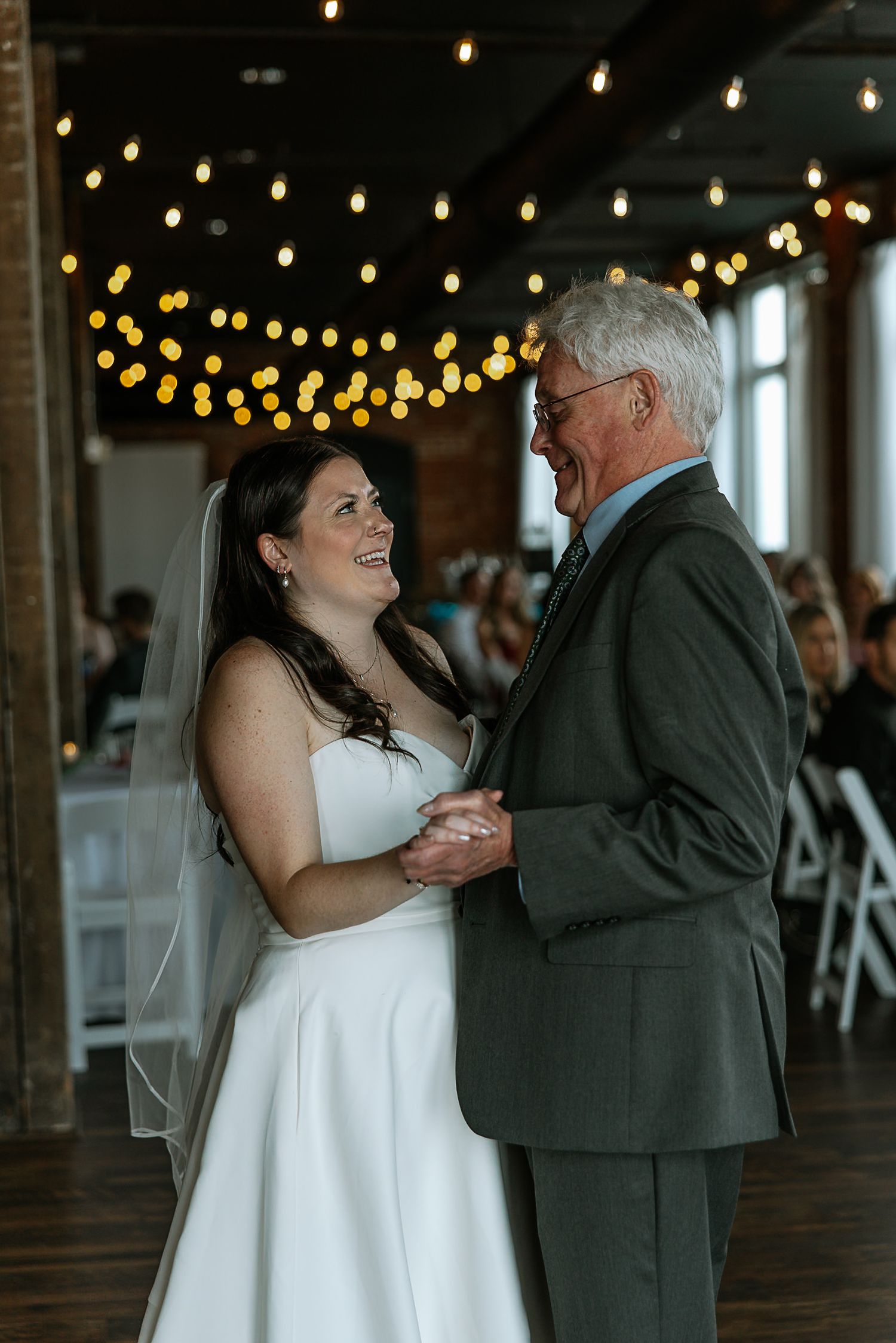 Bride shares a joyful father-daughter dance under warm string lights at an indoor wedding reception.