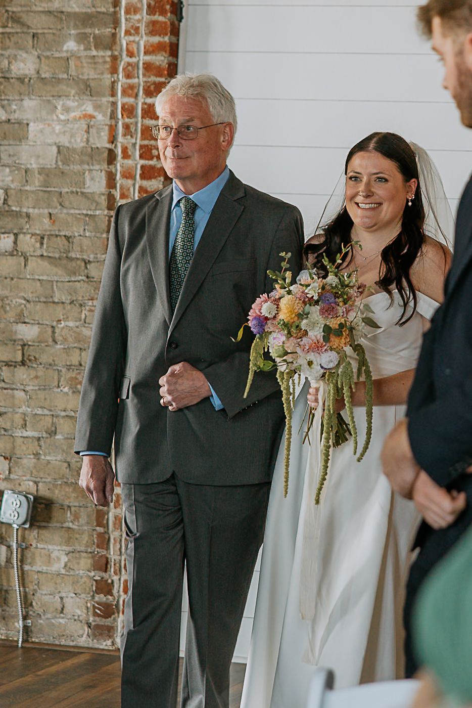 Bride with colorful bouquet walks down aisle with older gentleman in gray suit at wedding.