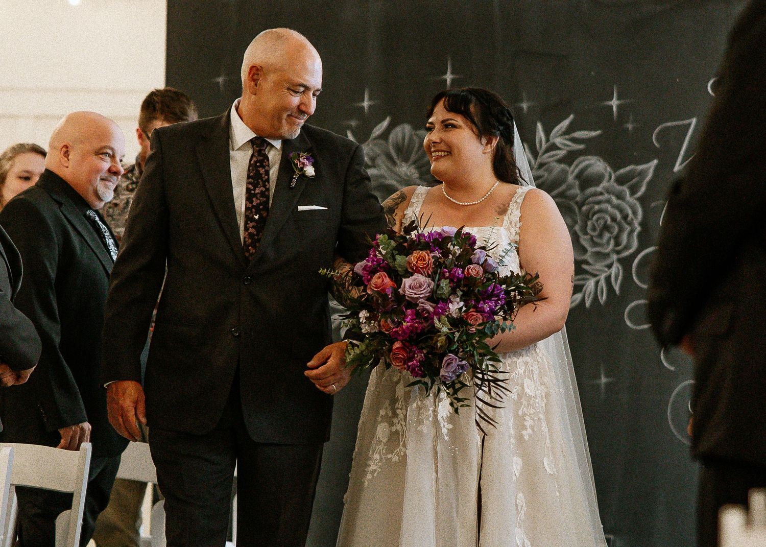 Smiling bride in lace gown walks down aisle with father before chalkboard backdrop.