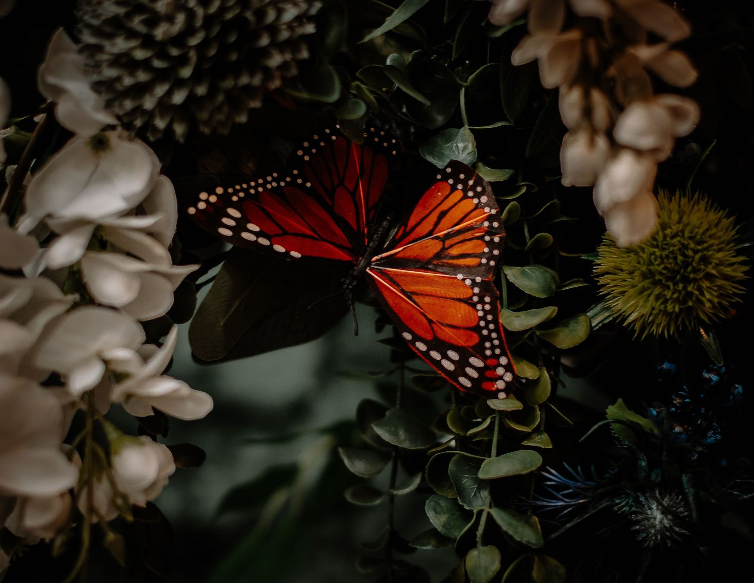A vibrant red monarch butterfly rests among lush green leaves and soft blooms in a dark, moody floral arrangement.