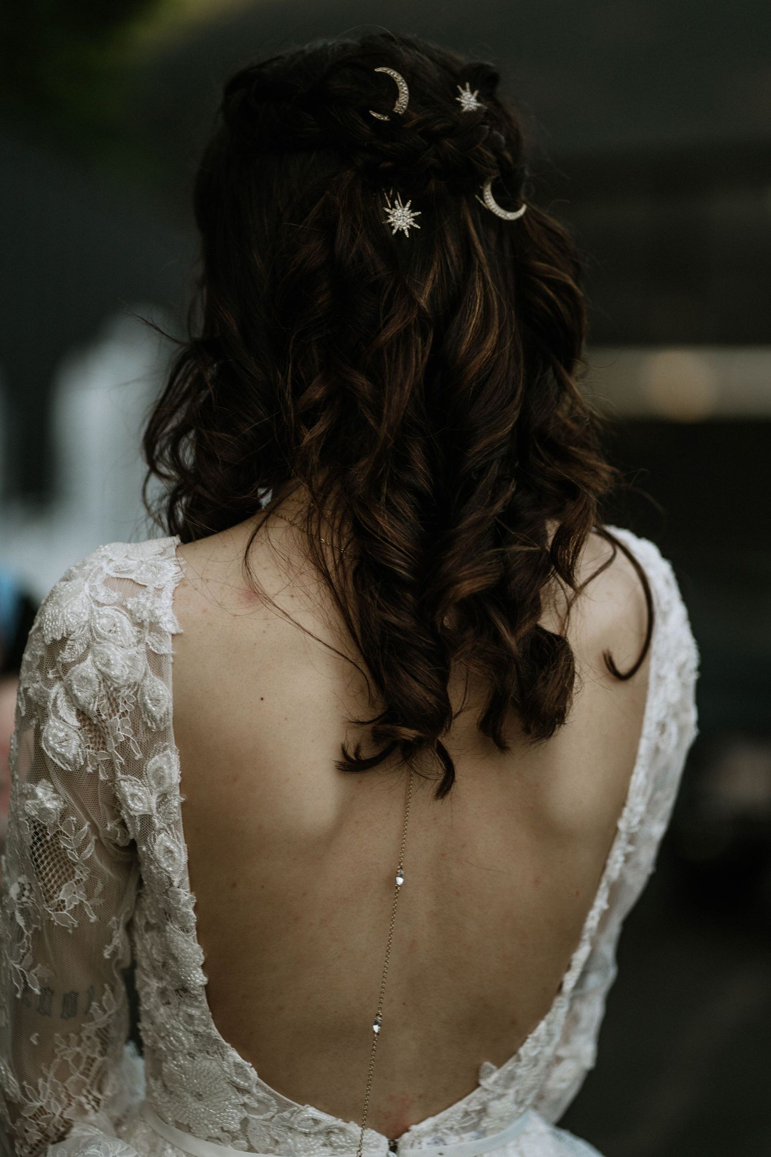 Bride with dark curly hair adorned with gold moon and star pins, wearing a backless lace wedding dress with delicate back necklace.