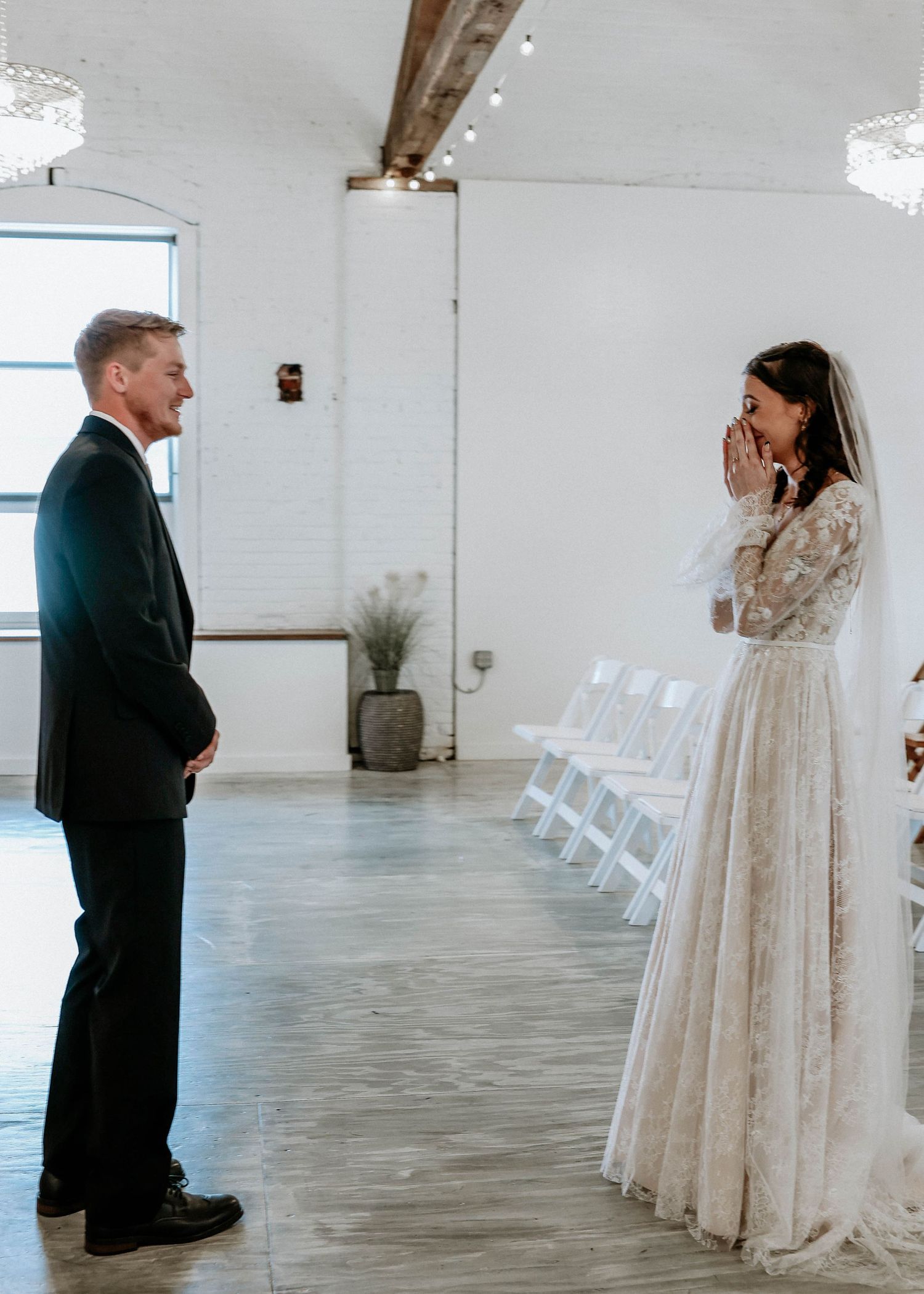 Bride in lace gown covers her mouth in emotional first look with groom in black suit at elegant white wedding venue.