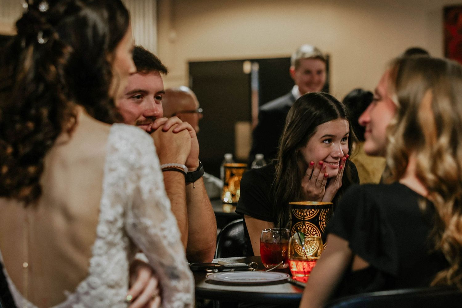 Guests laughing and celebrating at a wedding reception dinner table in a warmly lit venue.