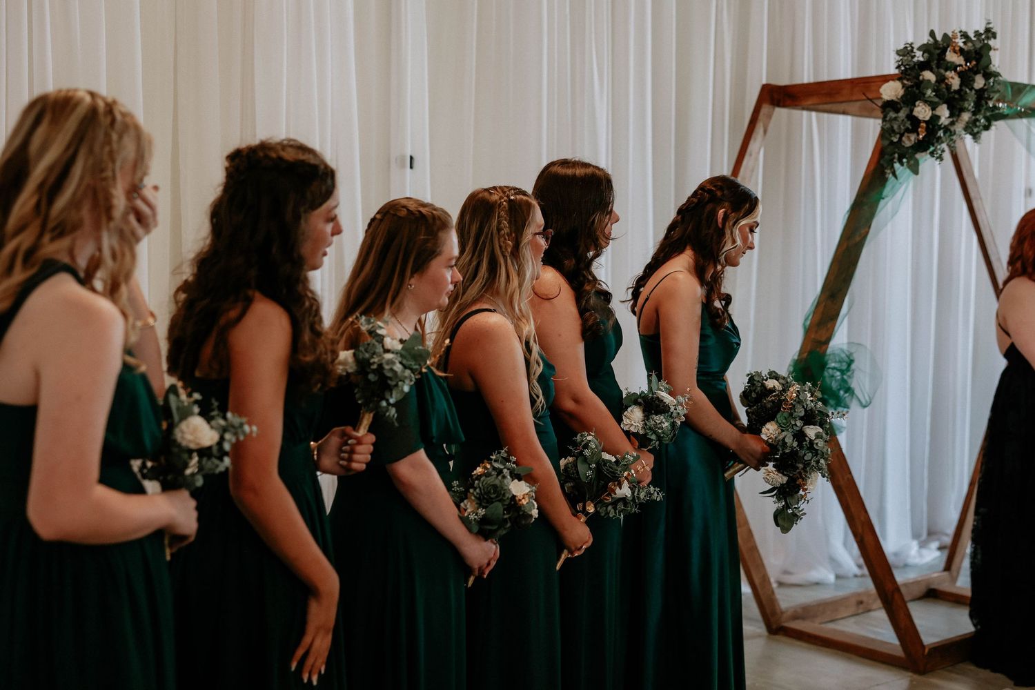Bridesmaids in emerald green gowns holding white bouquets stand in a row near a wooden arch at a wedding ceremony.