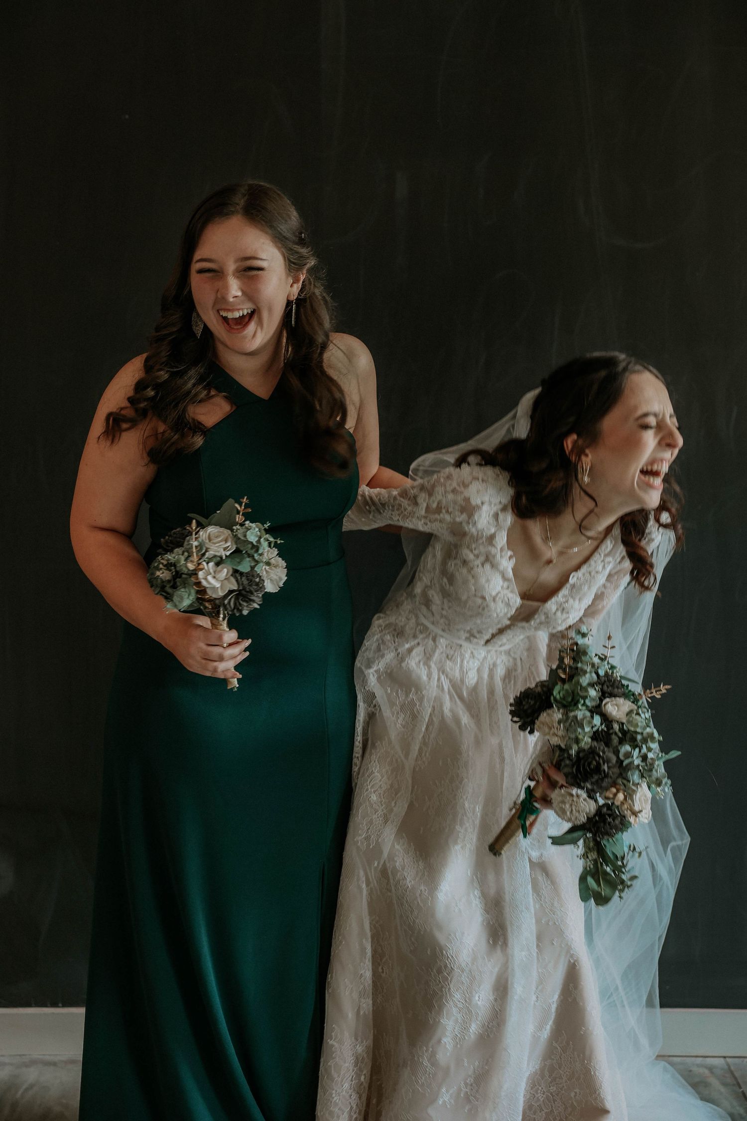 Laughing bridesmaid in emerald dress adjusts bride's veil while both hold floral bouquets indoors.