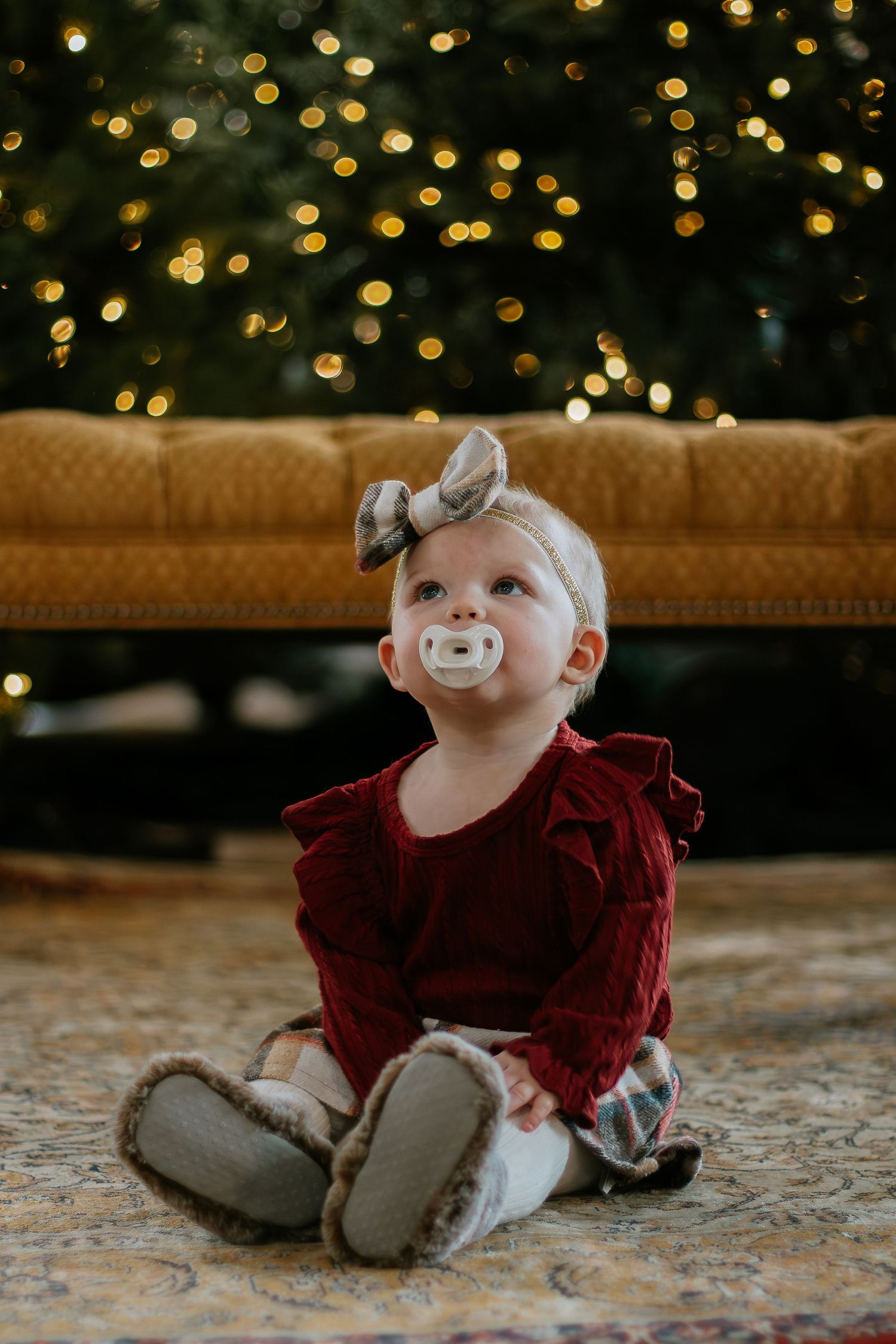 Adorable baby girl in red dress with bow headband sitting near Christmas tree at Mill Street Studios in Cincinnati, Ohio.