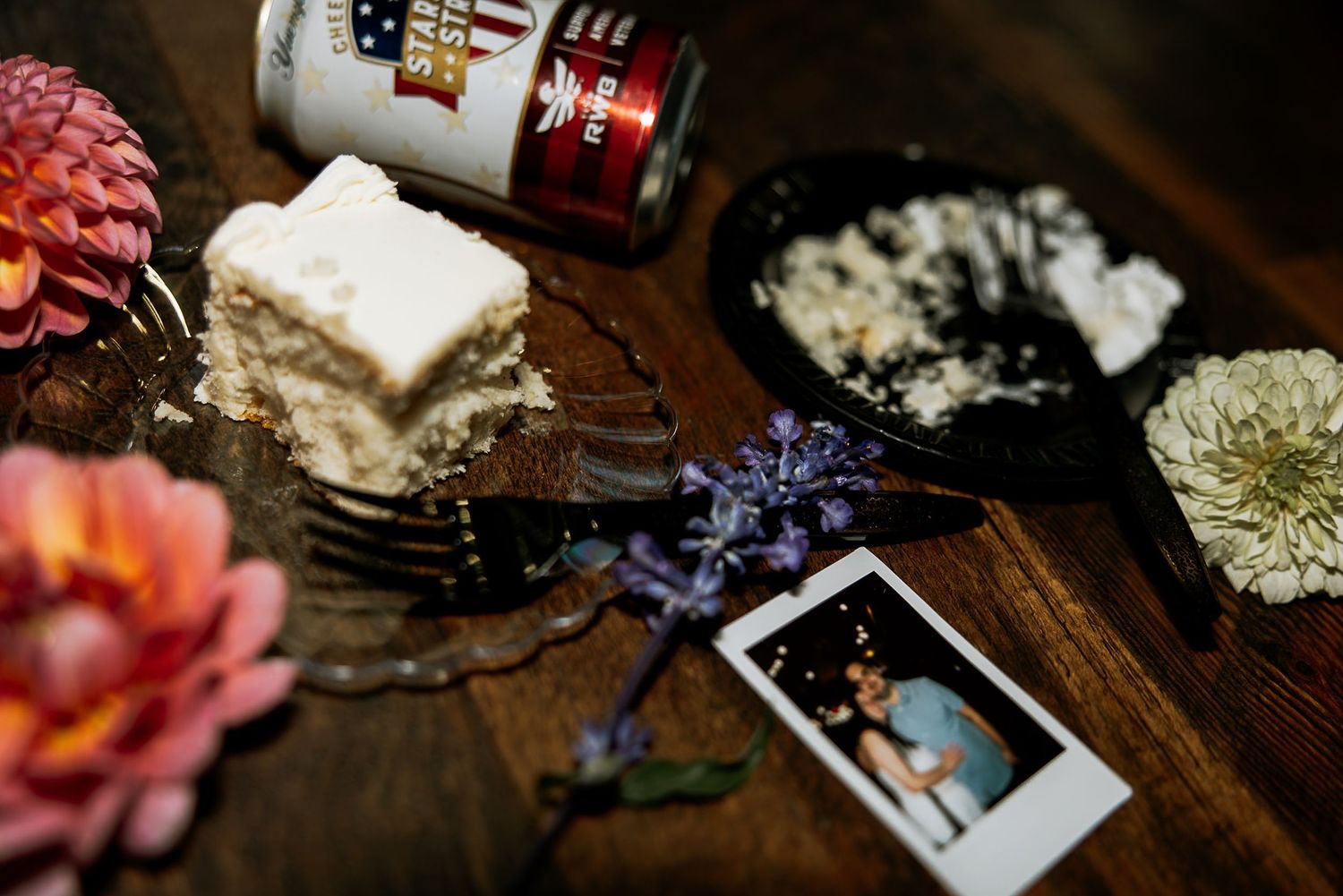 Rustic wedding flat lay with cake slice, Polaroid photo, flowers, and a beer bottle on wood.