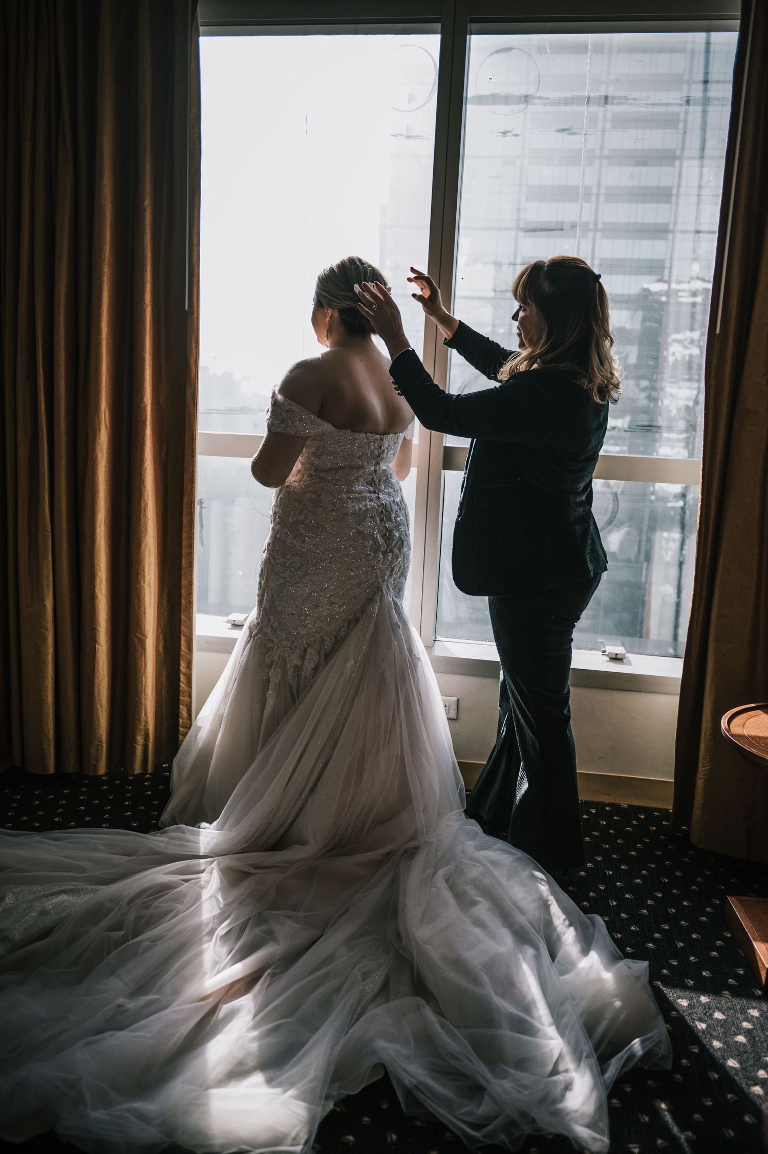Stylist adjusts bride's updo hairstyle as she stands in lace mermaid gown by hotel window.