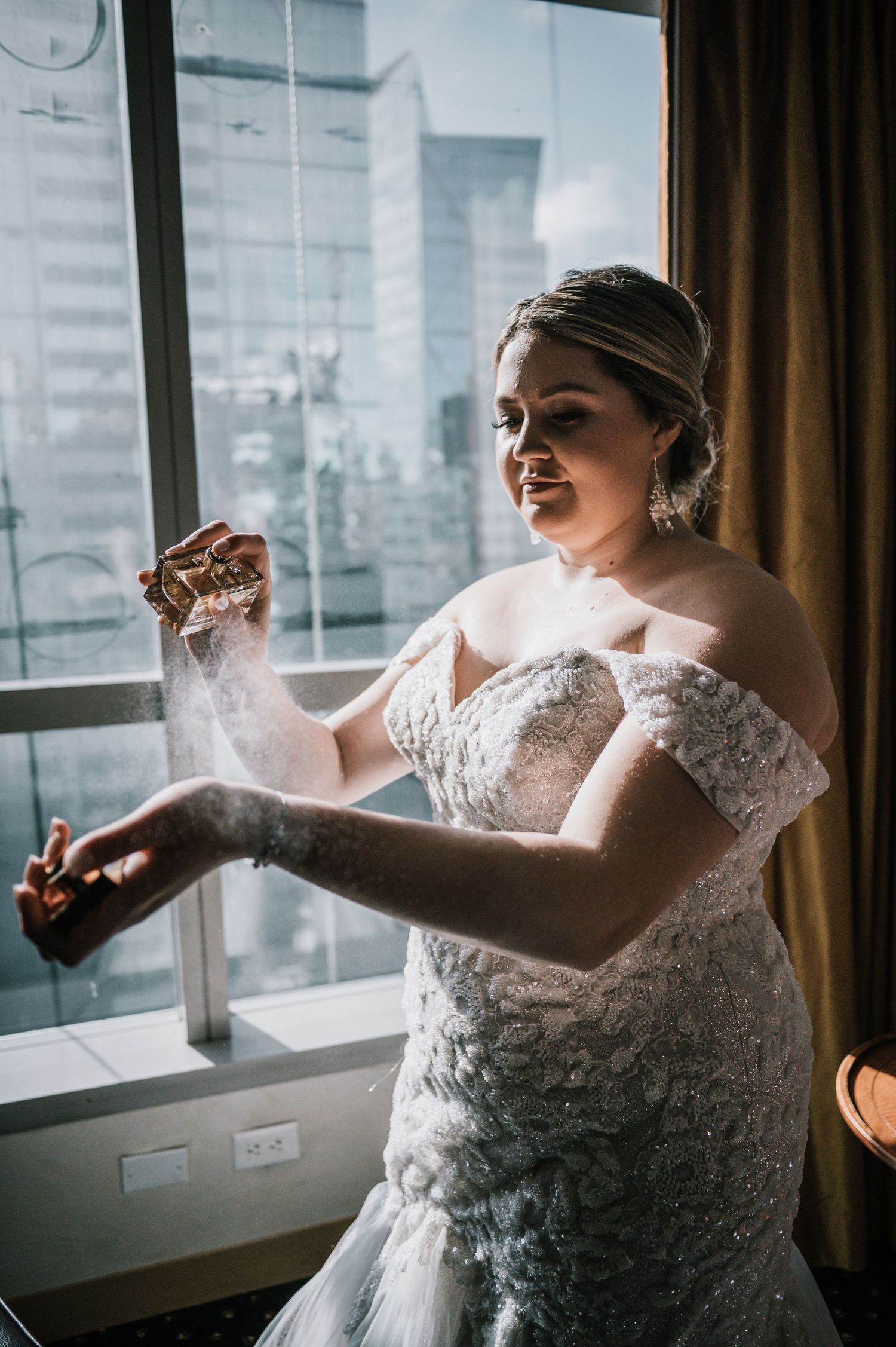 Bride in off-shoulder lace wedding gown spraying perfume by hotel window.