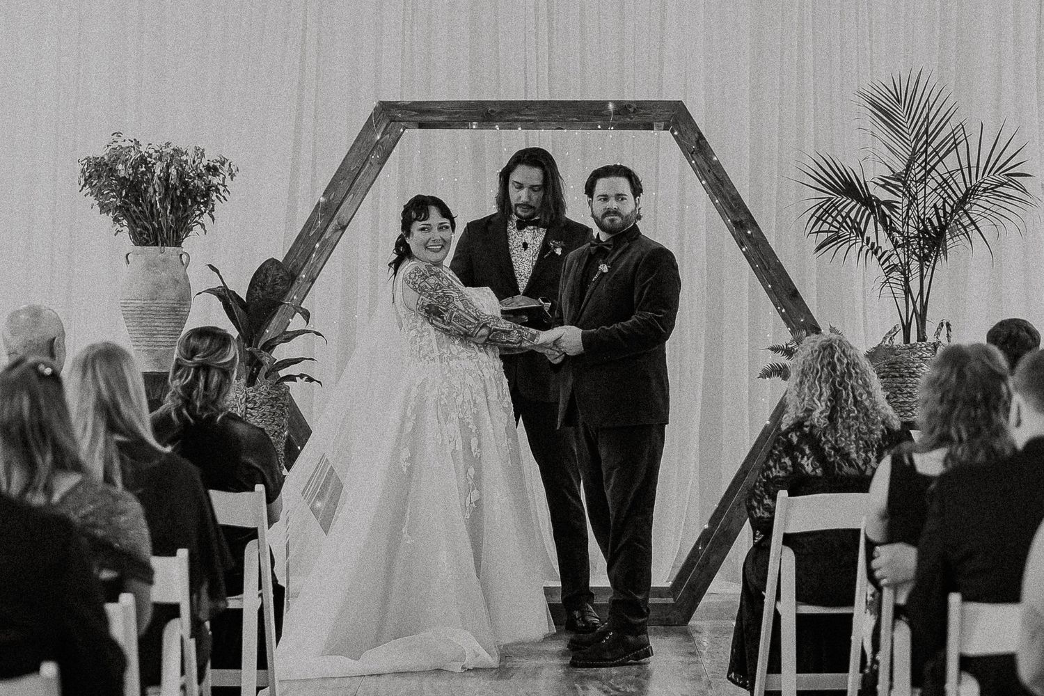 Black and white wide shot of bride and groom at hexagonal arch altar with seated guests.