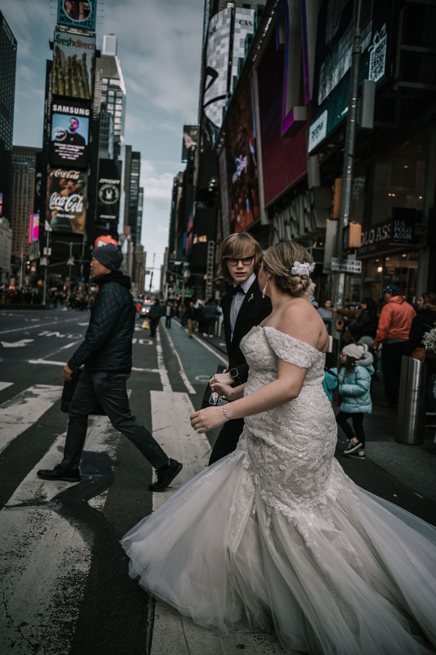 Bride and groom crossing busy Times Square street, bride in lace mermaid gown with long train.