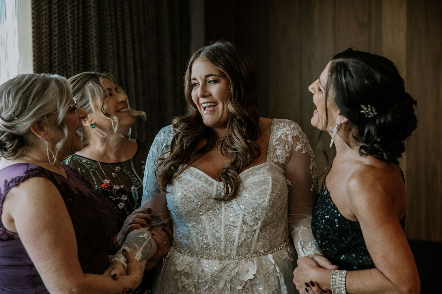 Bride in lace wedding gown laughing joyfully with bridesmaids during wedding preparations.