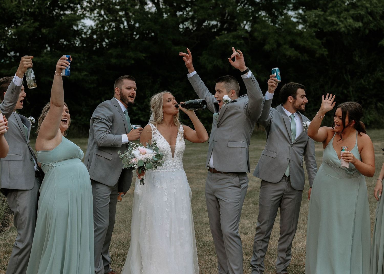 Wedding party celebrating outdoors, bride in white lace gown holding bouquet, groomsmen in gray suits, bridesmaids in sage green dresses.