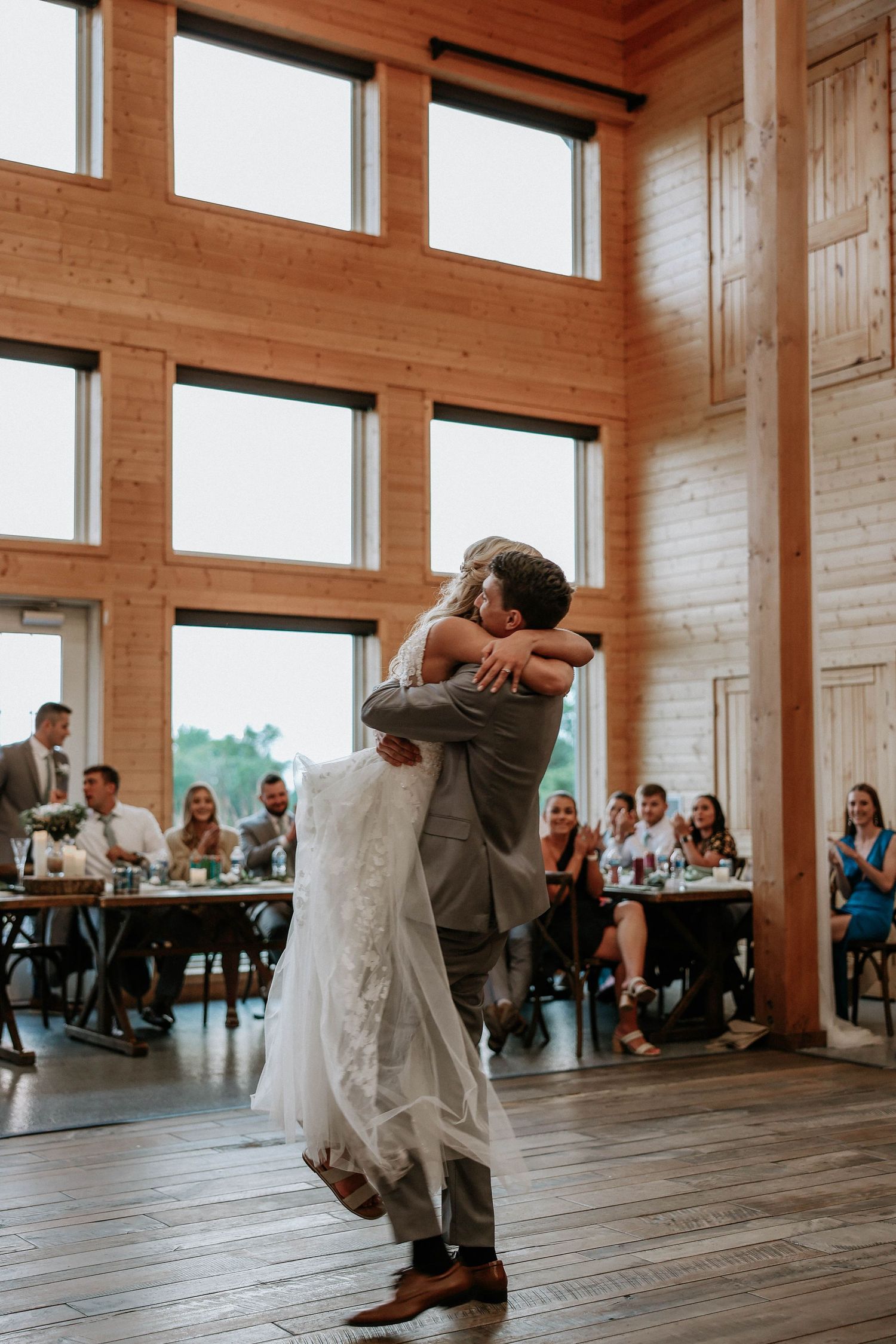 Groom lifts bride in lace wedding gown during first dance at rustic barn reception with guests.