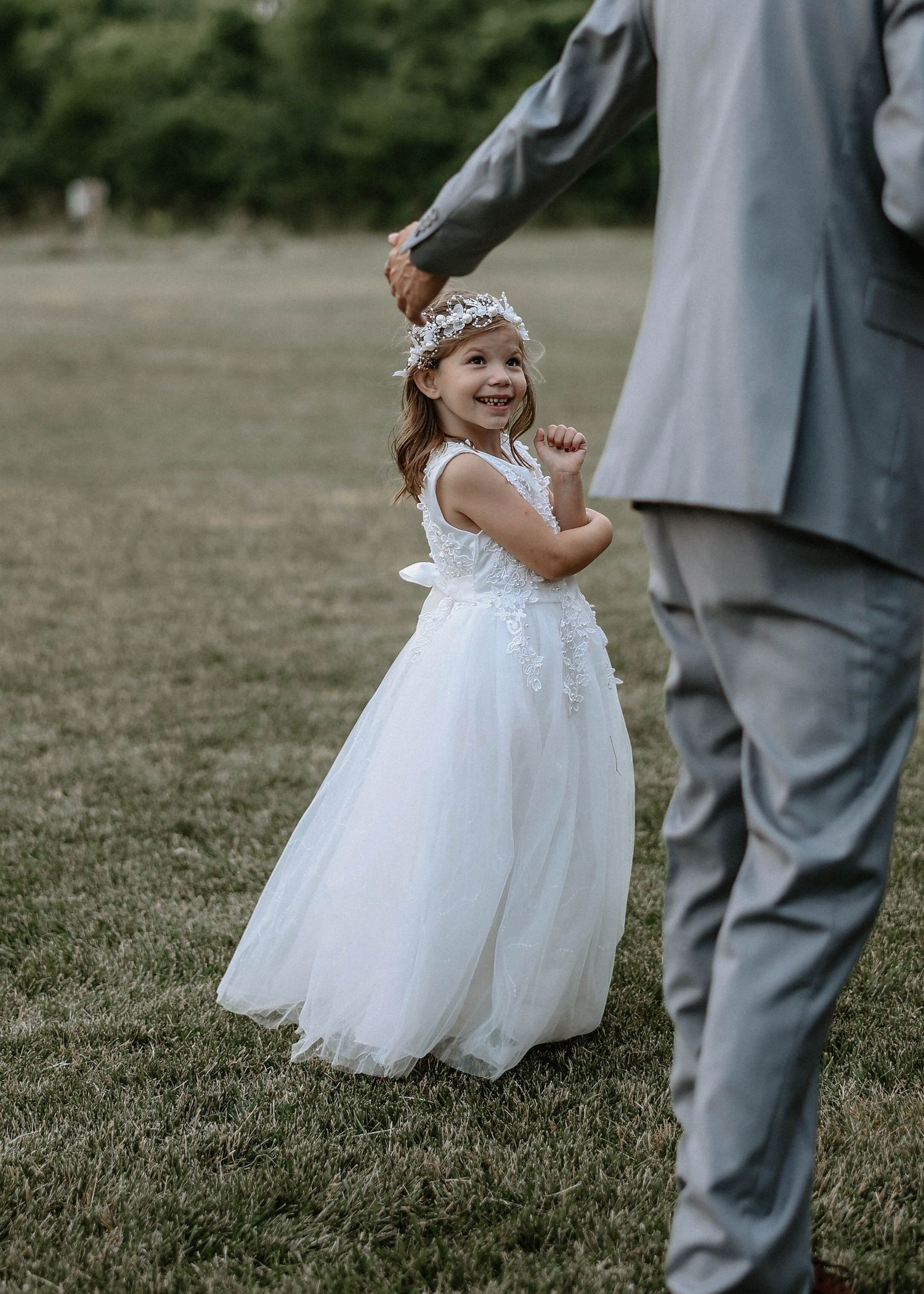 Smiling flower girl in white tulle dress with floral crown dancing with suited groomsman outdoors.