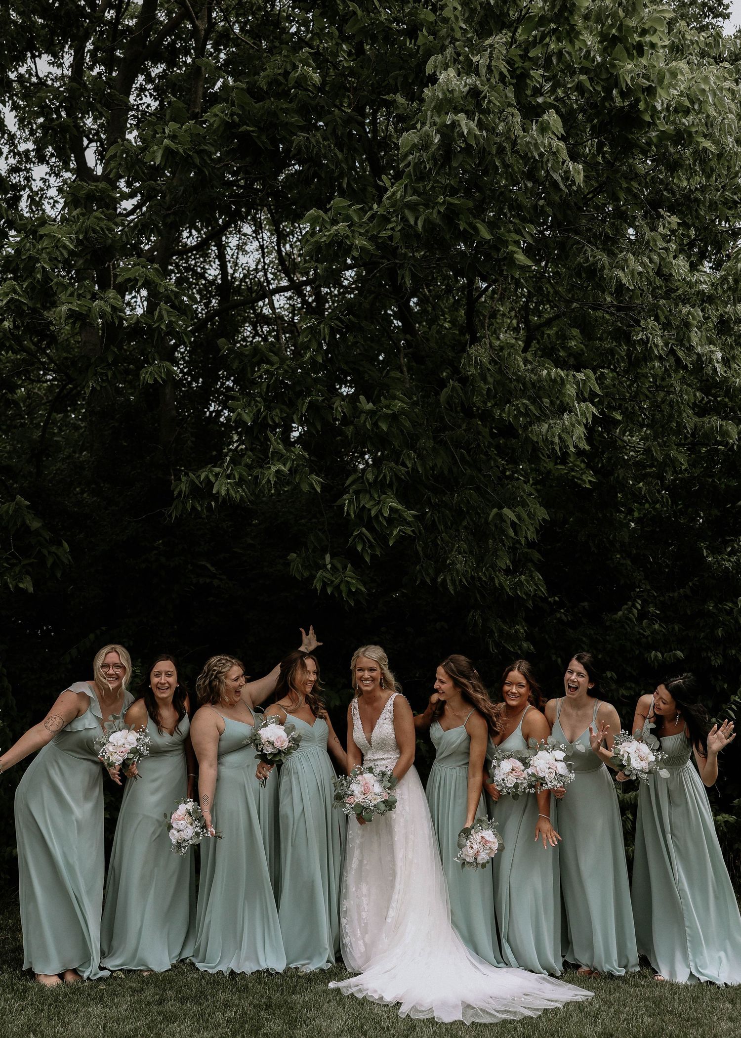 Bride in white gown laughing with eight bridesmaids in sage green dresses outdoors.