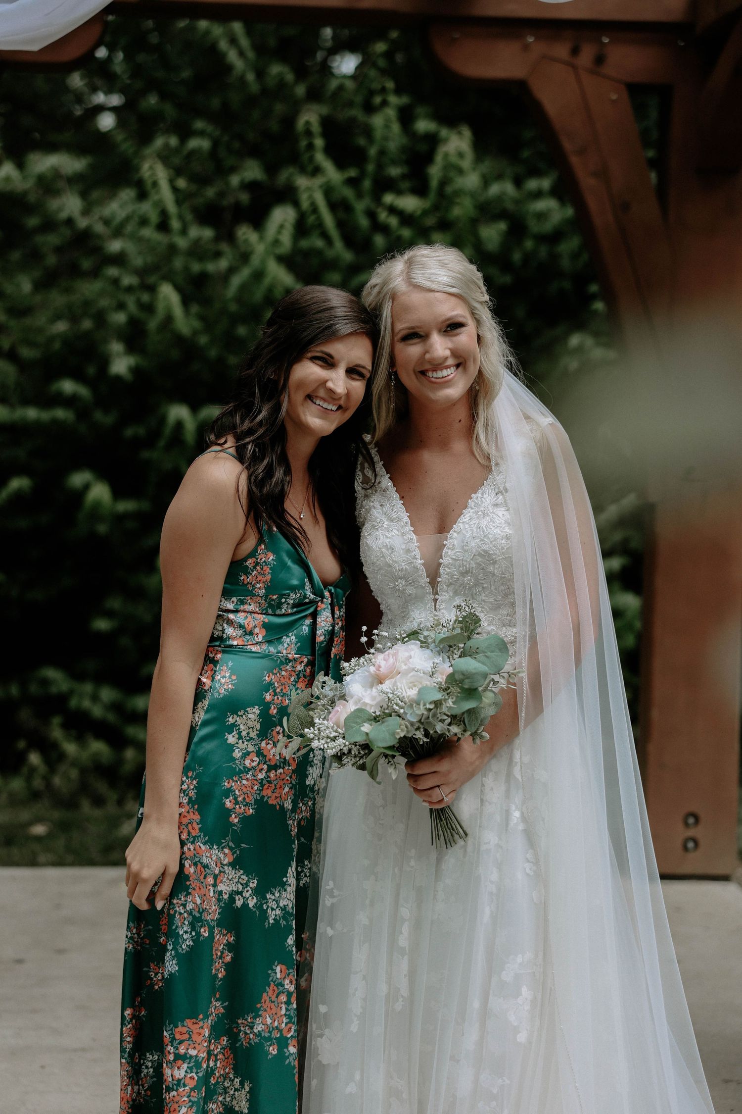 Bride in lace wedding gown with veil holding bouquet posing with guest in green floral dress outdoors.