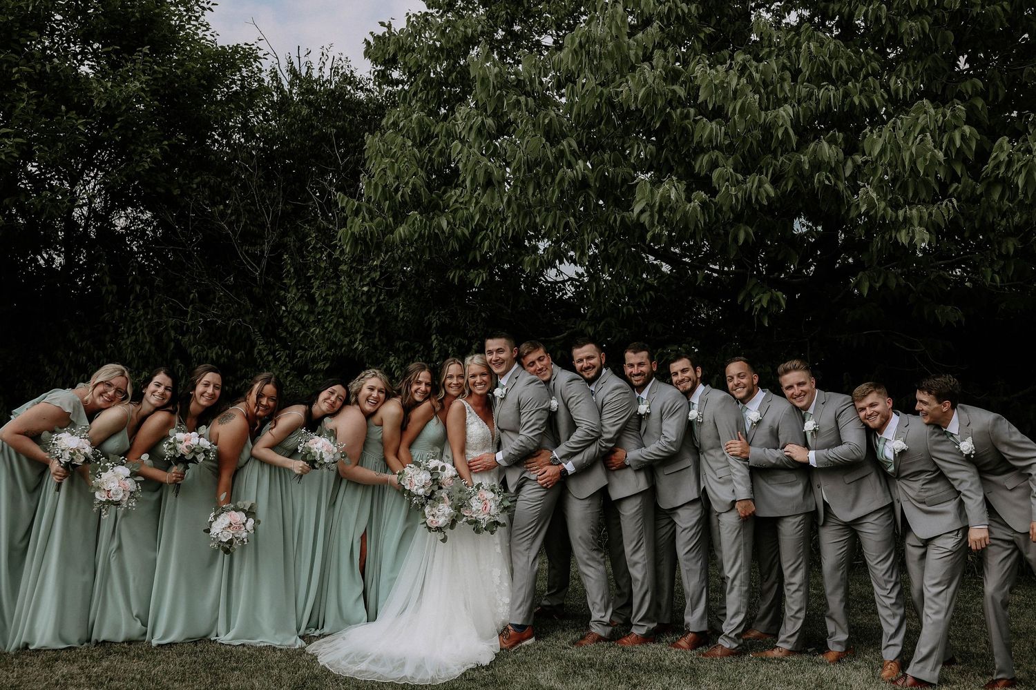 Wedding party poses outdoors, bridesmaids in mint green gowns and groomsmen in gray suits flanking bride and groom.