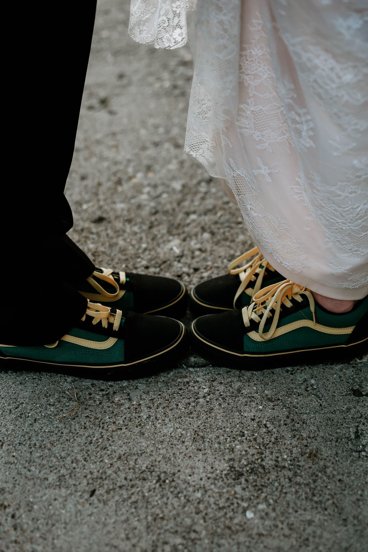 Bride and groom wearing matching green and gold Vans sneakers on their wedding day, standing on gray pavement.