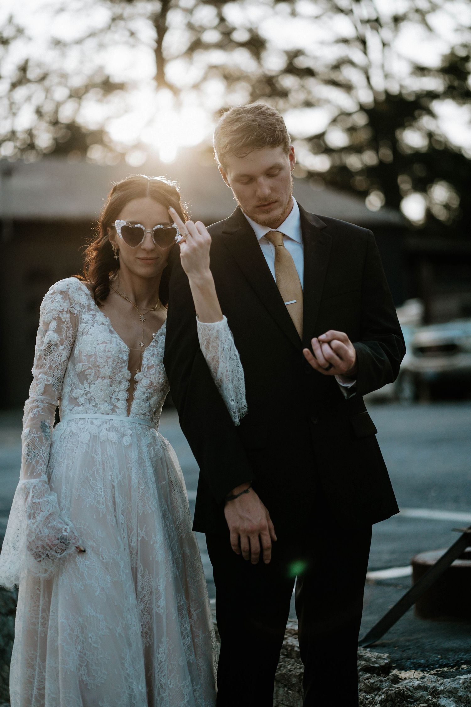 Bride in lace gown with heart sunglasses stands beside groom in black suit and gold tie at golden hour outdoor wedding.