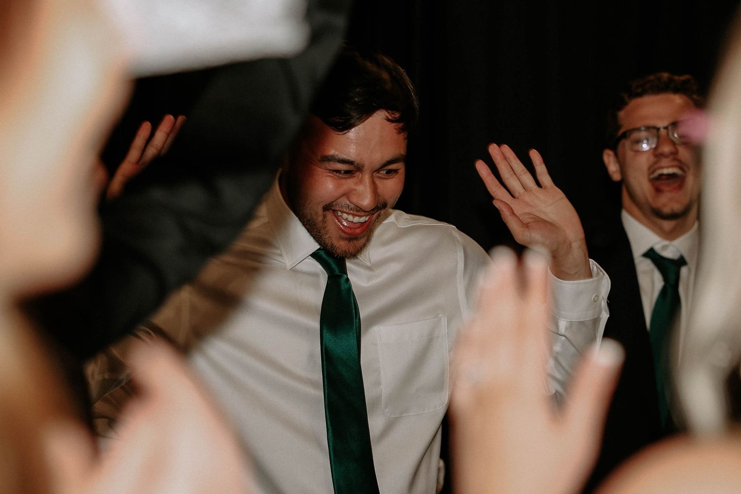 Two men in white shirts and green ties celebrate joyfully on a wedding dance floor with hands raised.