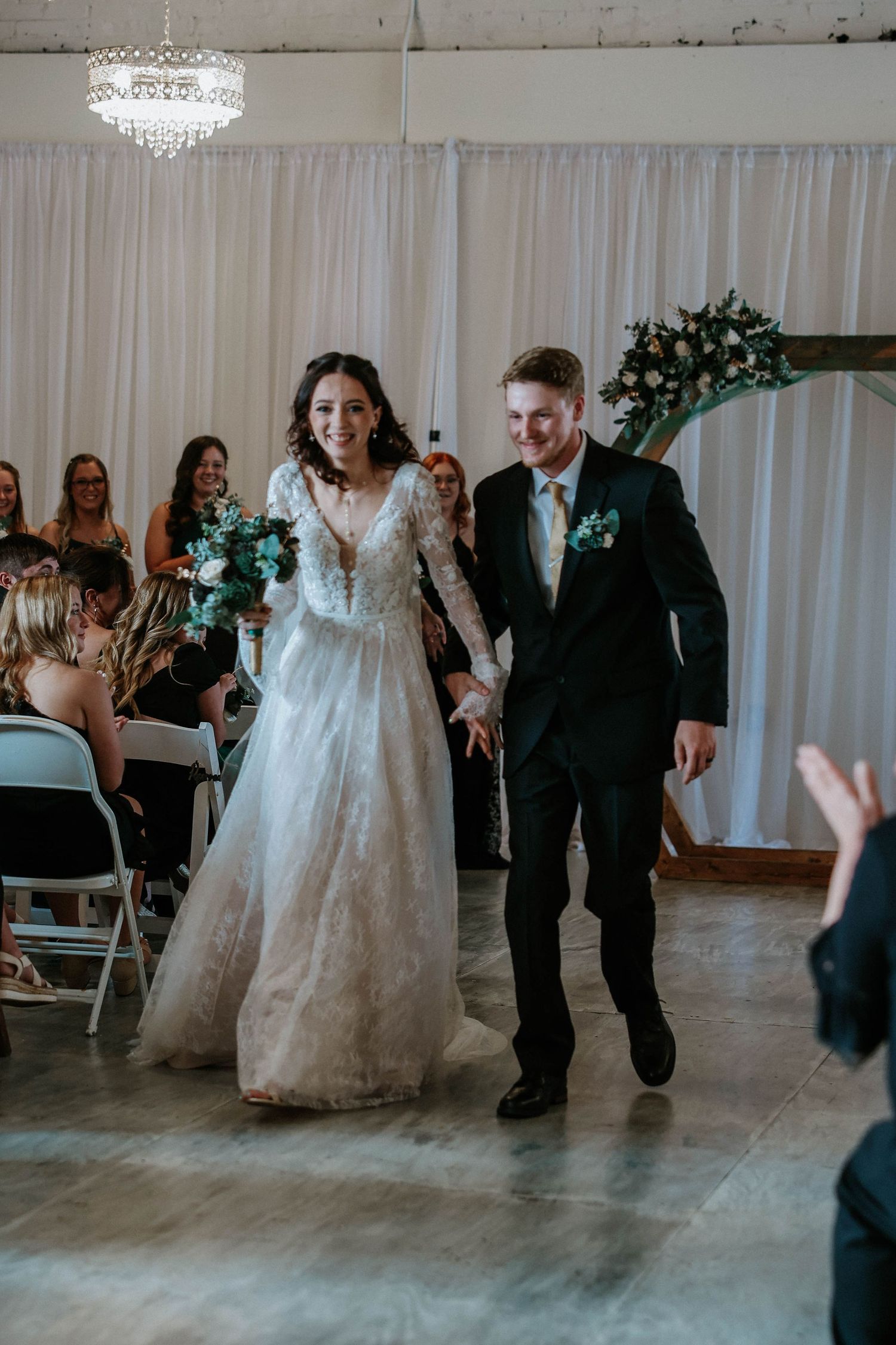 Bride in lace gown and groom in black suit walk joyfully down the aisle after wedding ceremony indoors.