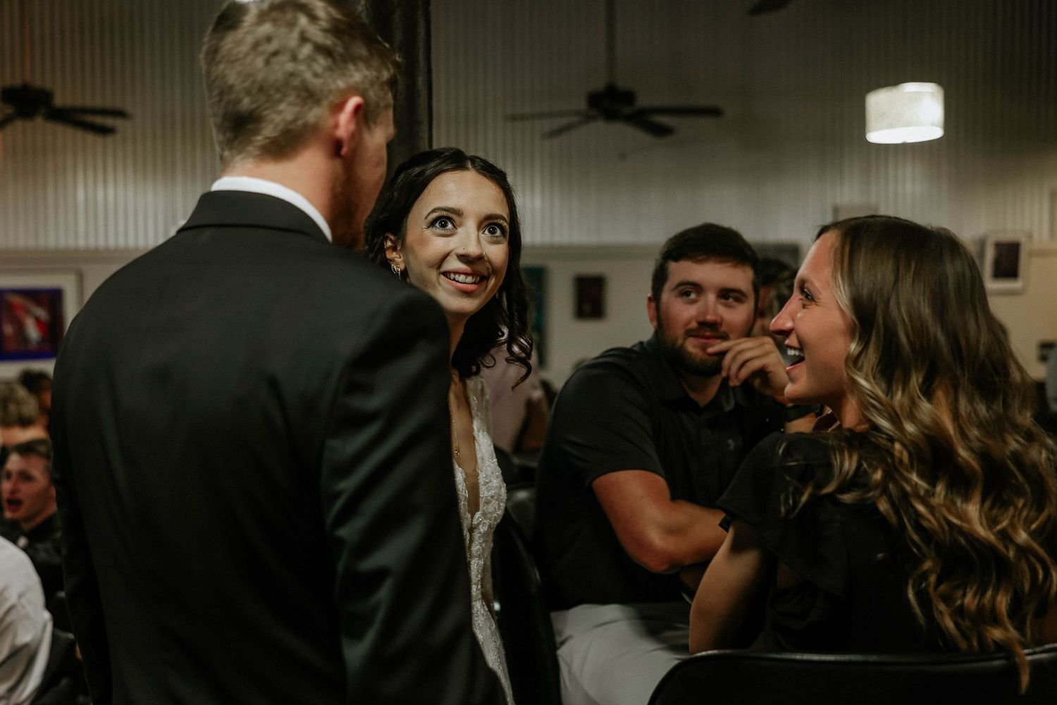 Bride smiling at groom during wedding reception while guests laugh and celebrate in background.