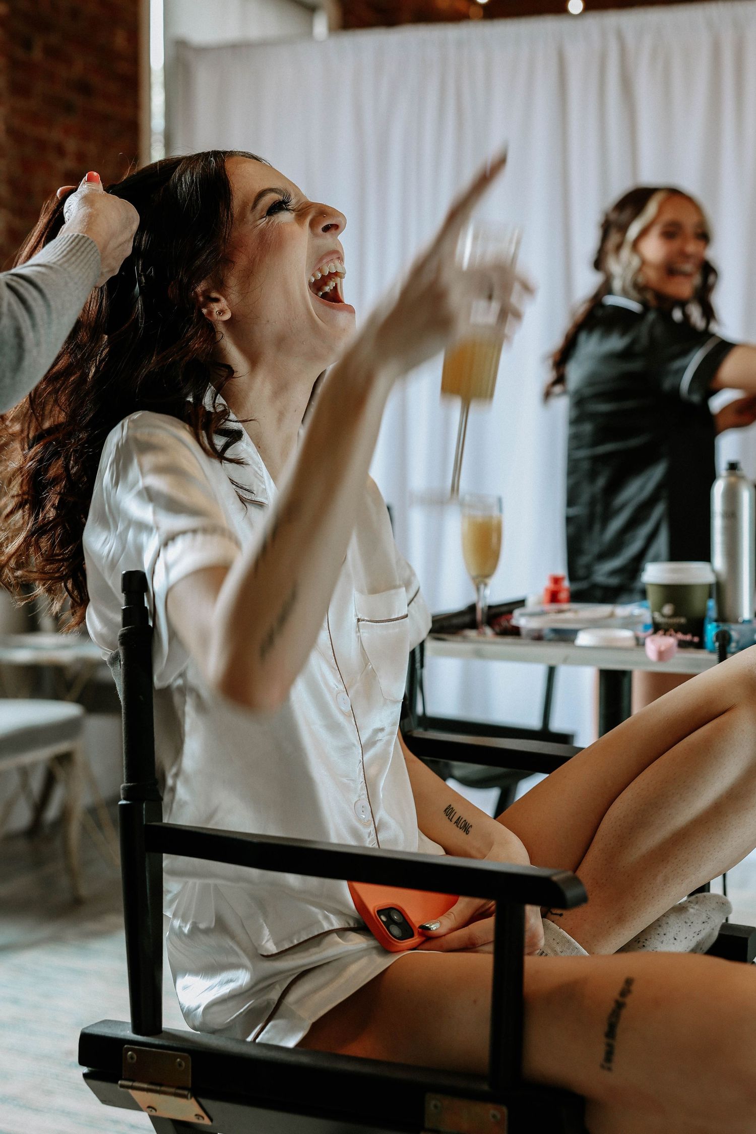 A laughing woman in white pajamas holds a mimosa glass while sitting in a director's chair during a bridal getting-ready session.