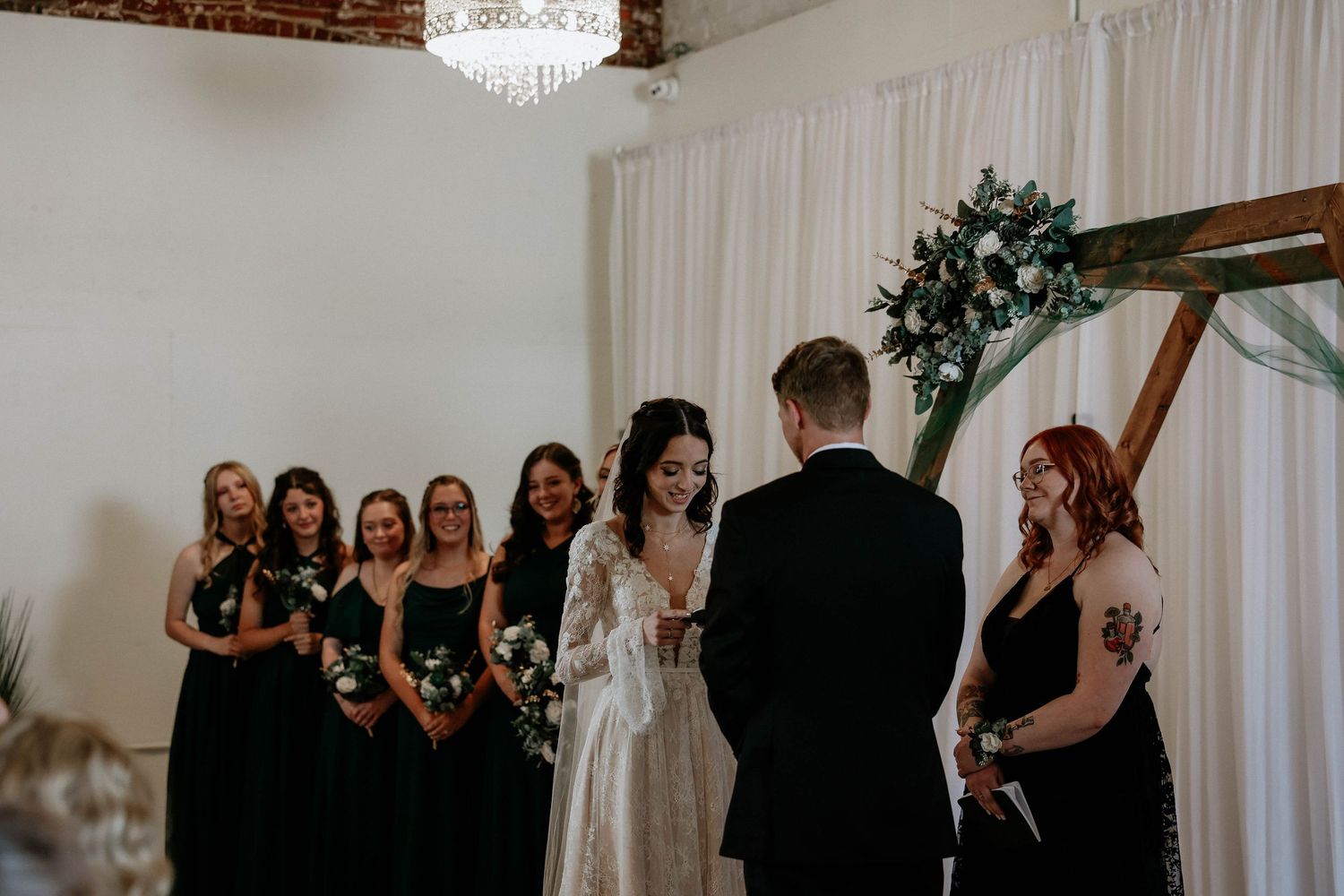 Bride and groom exchange vows at altar with bridesmaids in black dresses standing beside wooden arch with greenery.