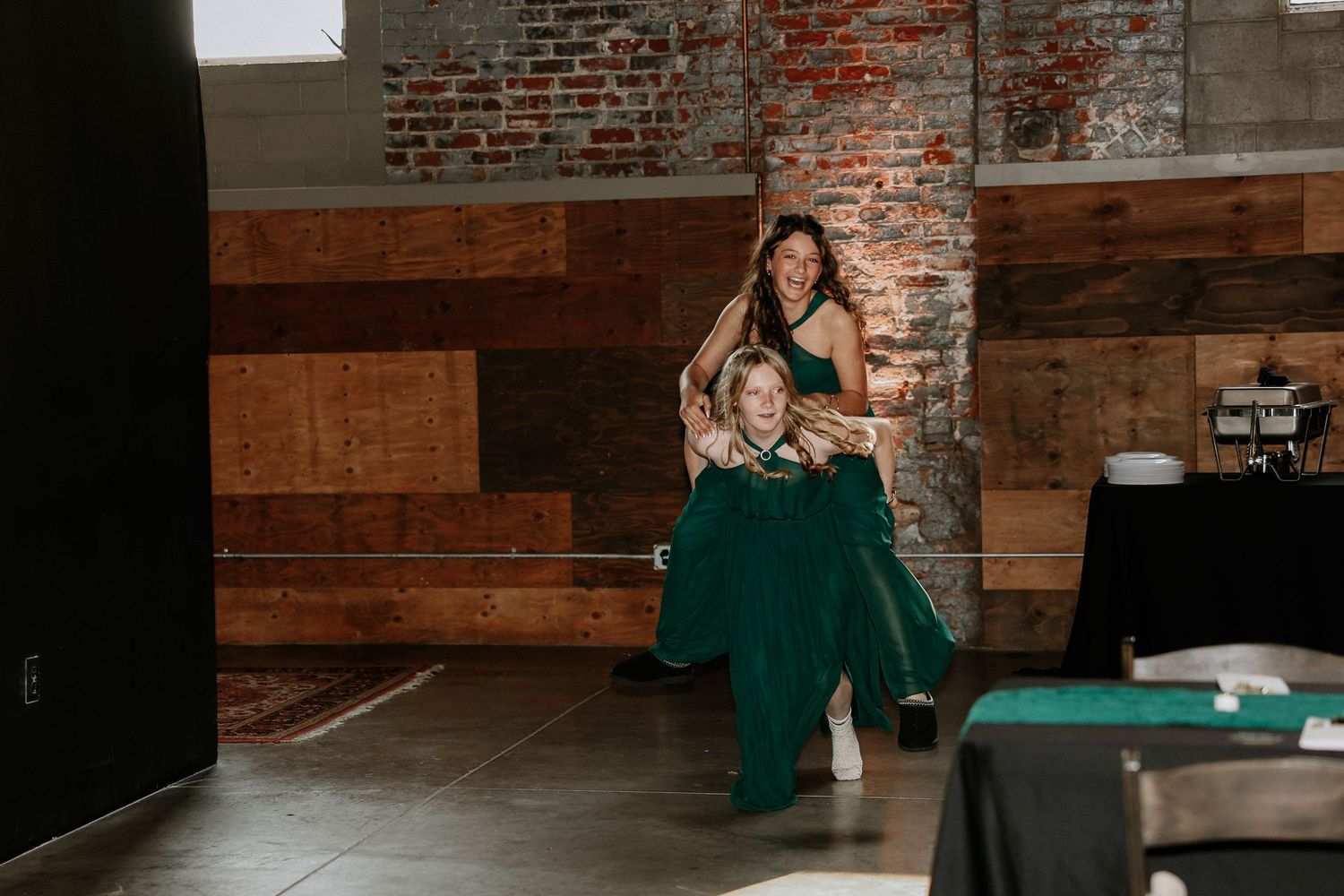 Two women in emerald green gowns laugh together at an indoor wedding reception venue with rustic brick walls.