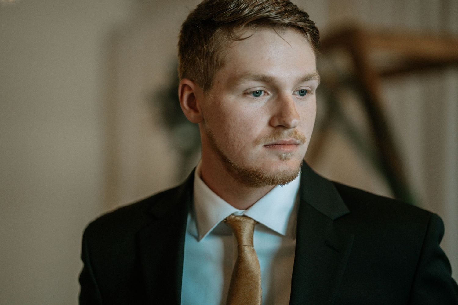 A young man in a dark suit and gold tie gazes thoughtfully to the side in a softly lit indoor setting.