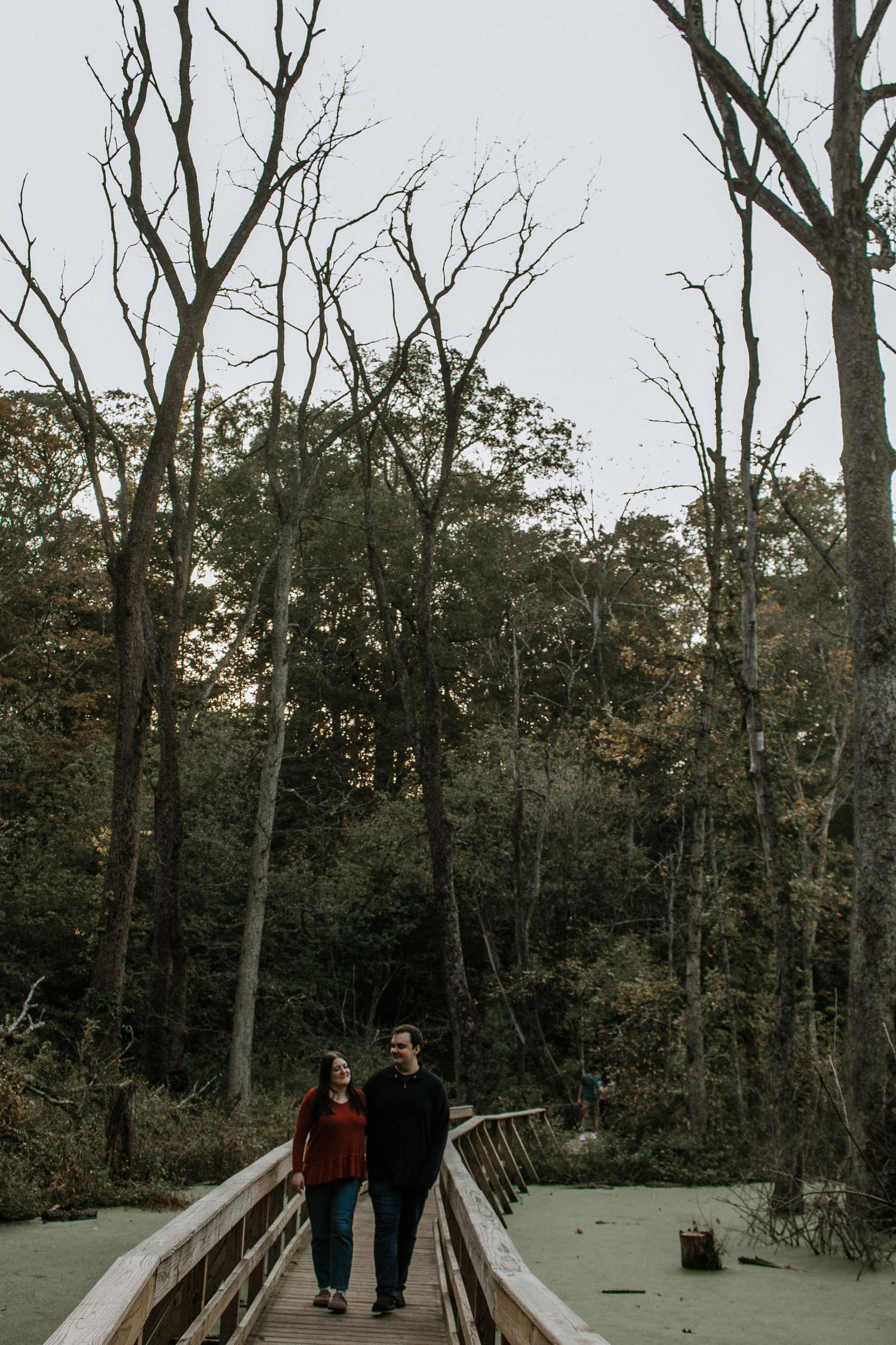 Couple holding hands on a wooden boardwalk bridge surrounded by tall bare trees in a forest in Yellow Springs, Ohio