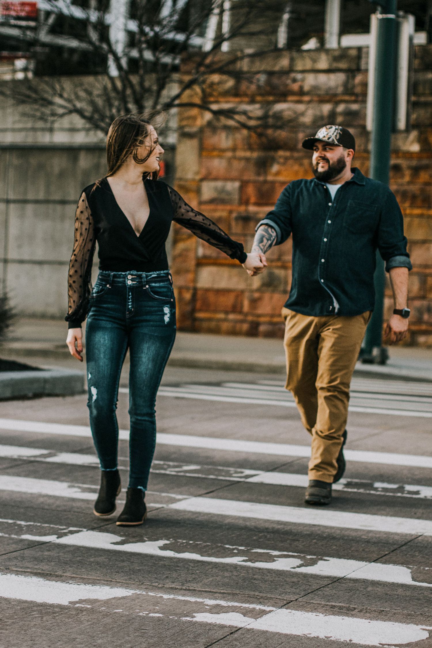 Stylish couple holding hands while crossing a city crosswalk, man with tattoos wearing a cap in Cincinnati, Ohio