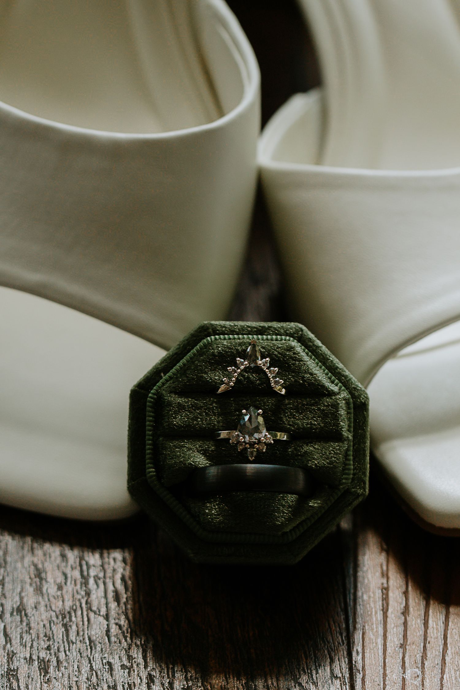 Green velvet ring box with wedding bands and engagement ring displayed in front of white bridal heels on wood at The Remedy in Dayton, Ohio