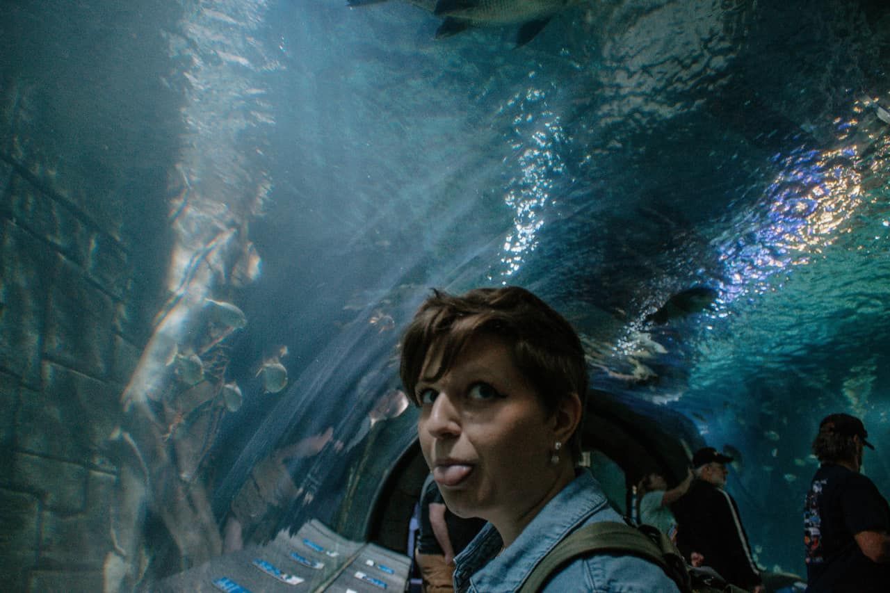 Woman makes a surprised face while standing in an underwater aquarium tunnel with rays of light above.