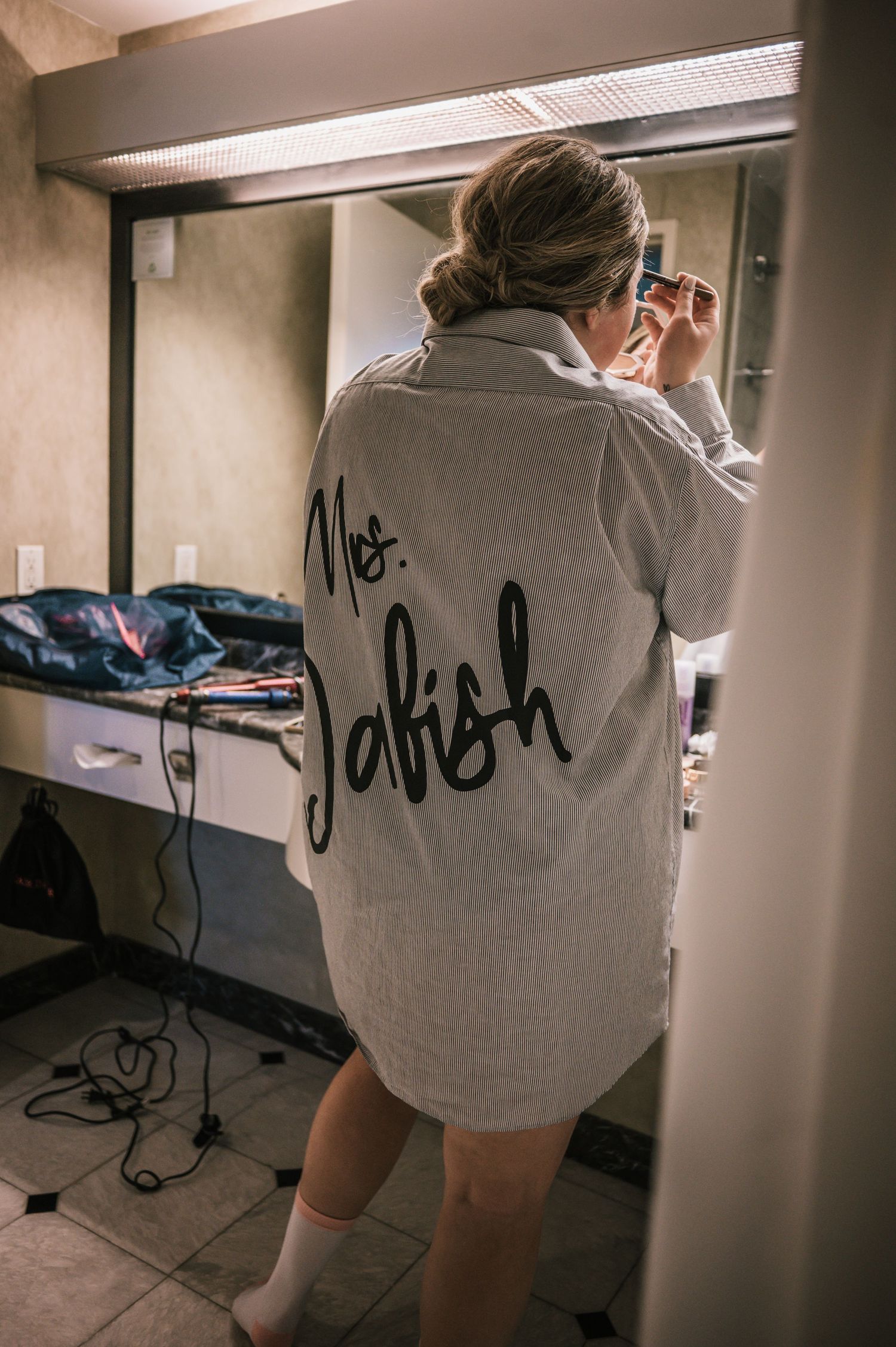 Bride wearing personalized Mrs. Jabish robe applying makeup in hotel mirror.